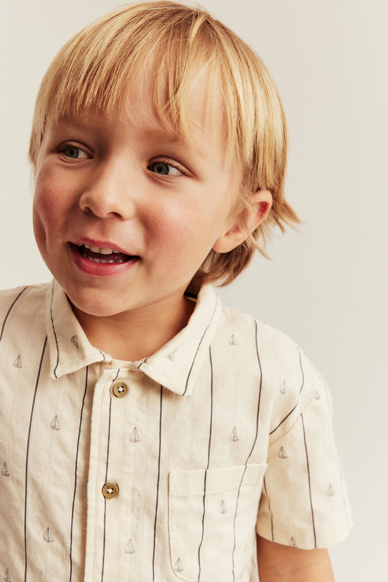 Nærbilde av barn i kremhvit stripete bomullsskjorte med seilbåtmønster og brune knapper.