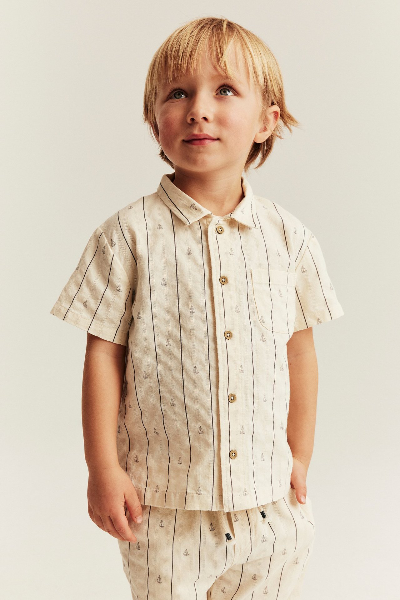 Widok z przodu: Dziecko w kremowej, prążkowanej koszuli bawełnianej z krótkim rękawem z żaglówkami, pasujące spodnie.