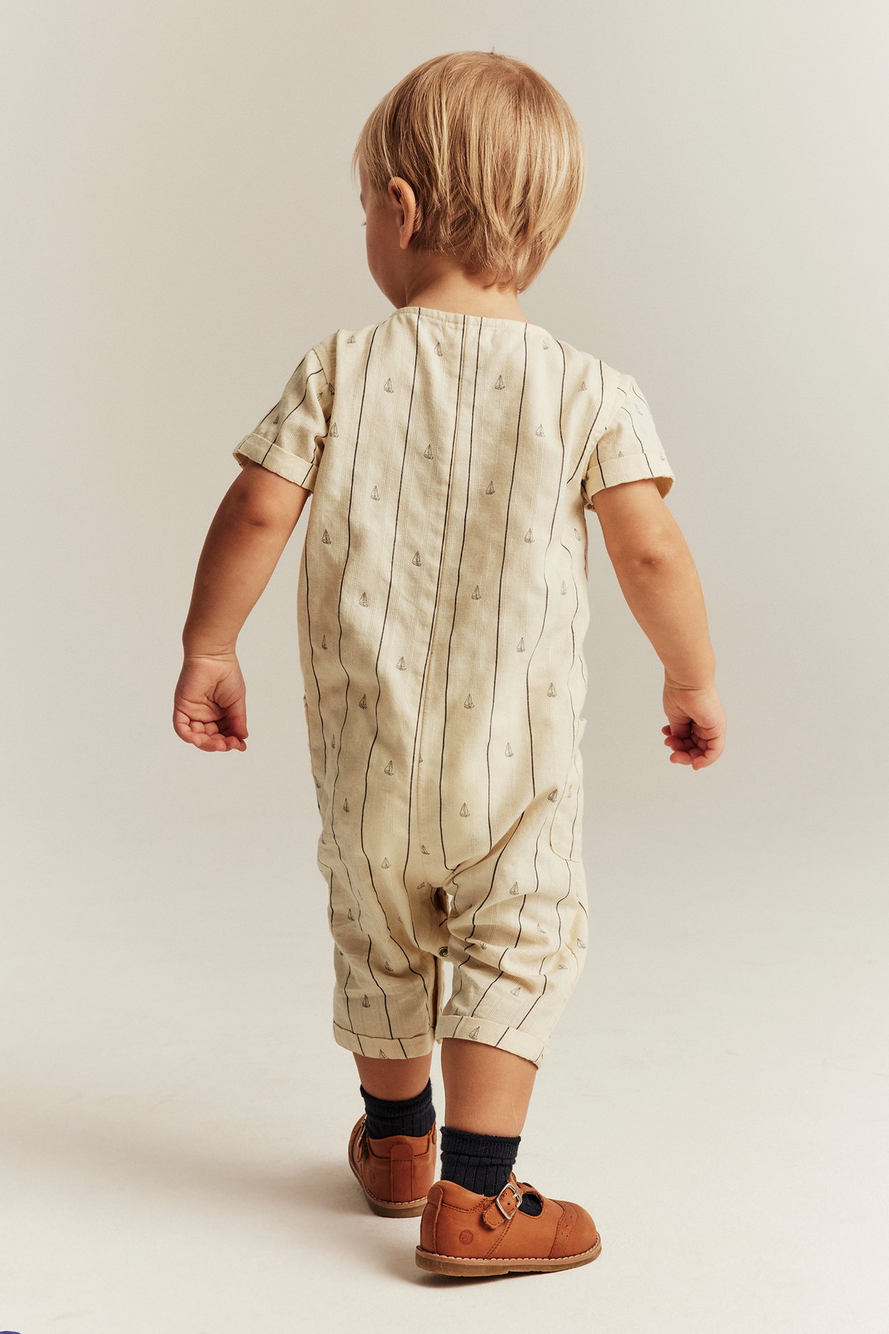 Bakfra av baby i naturhvit stripete bomulls-jumpsuit med seilbåter, mørke sokker og brune sko.