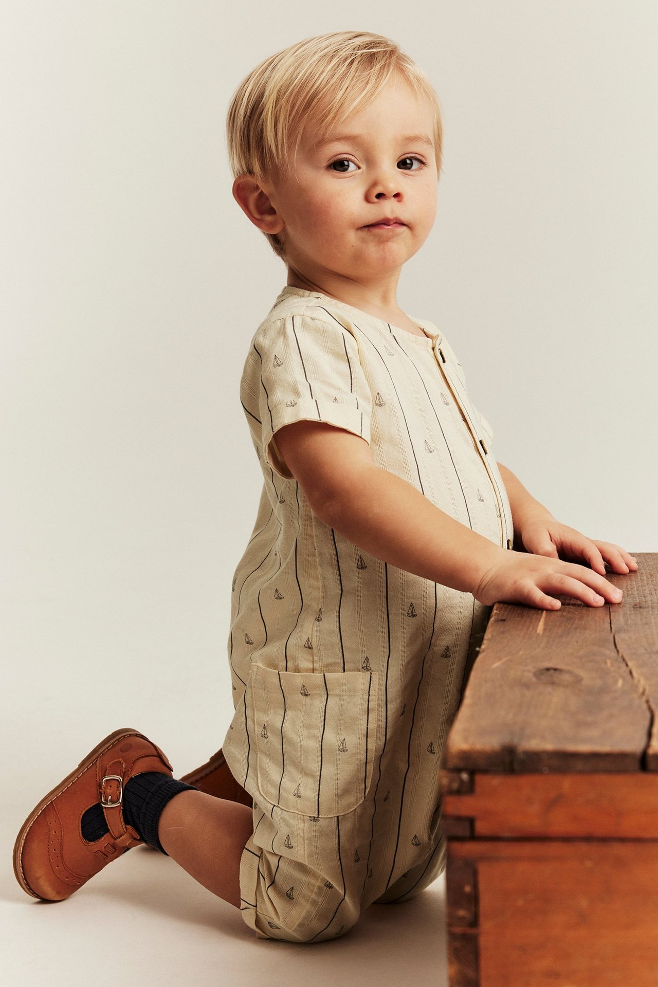 Sivukuva: vauva yllään vaaleanbeigessä lyhythihaisessa haalarissa purjevenekuvioilla, tummat sukat ja ruskeat kengät.