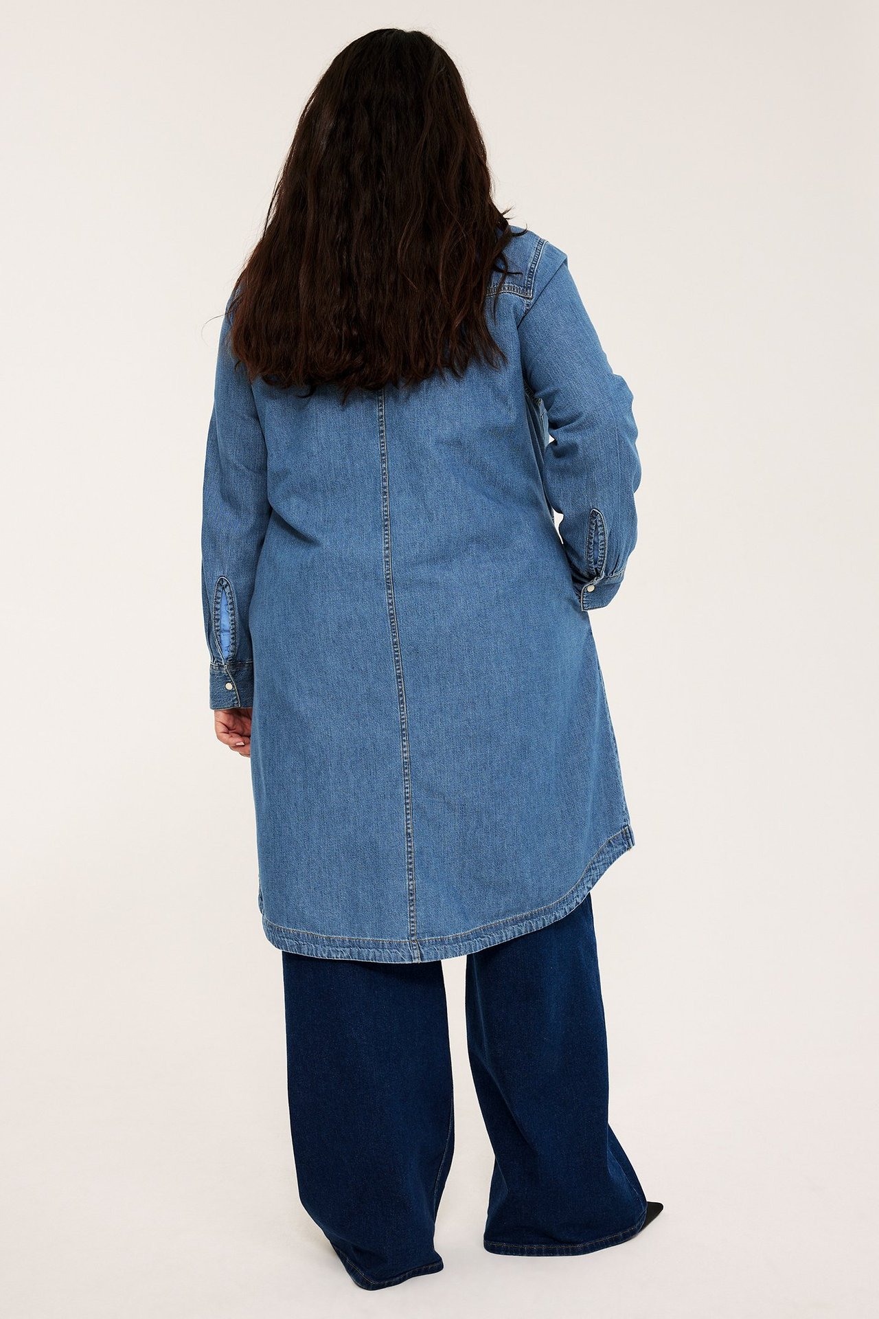 Ryggvy av en kvinna i ljusblå denim skjortklänning, buren över mörkblå vida jeans.