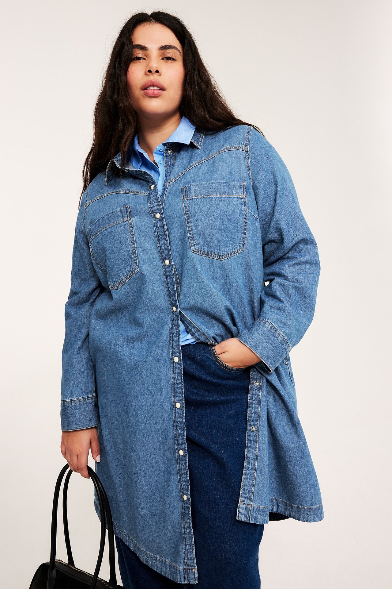 Front view: Kvinna i ljusblå jeansskjortklänning, blå topp, mörkblå kjol och svart handväska.