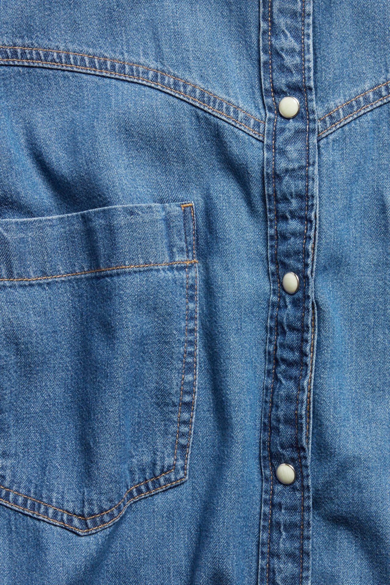 Zbliżenie na niebieską dżinsową sukienkę koszulową z kieszenią, szwami i białymi zatrzaskami.