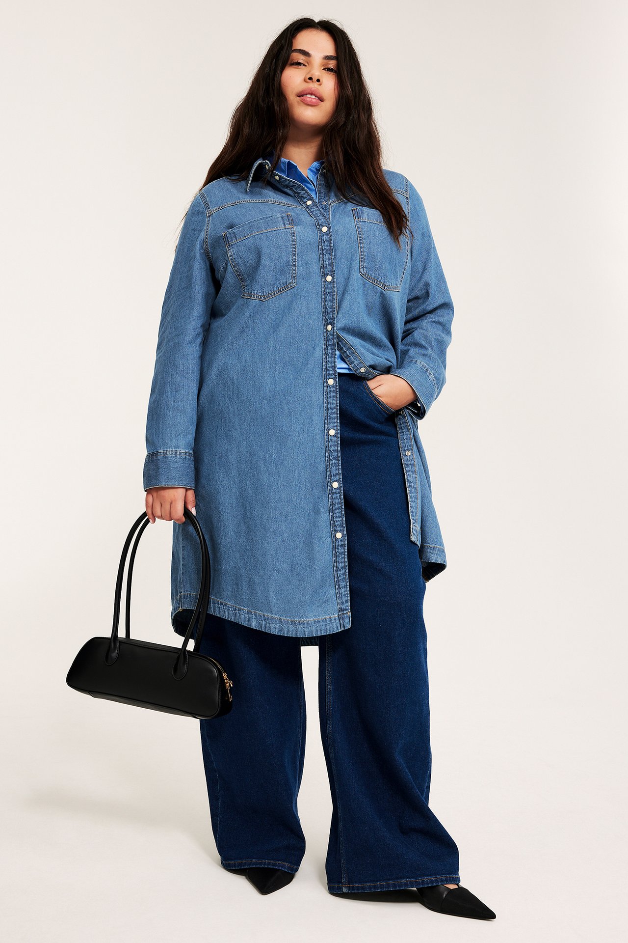 Framifrån: kvinna i öppen ljusblå jeansklänning, mörkblå jeans och håller en svart handväska.