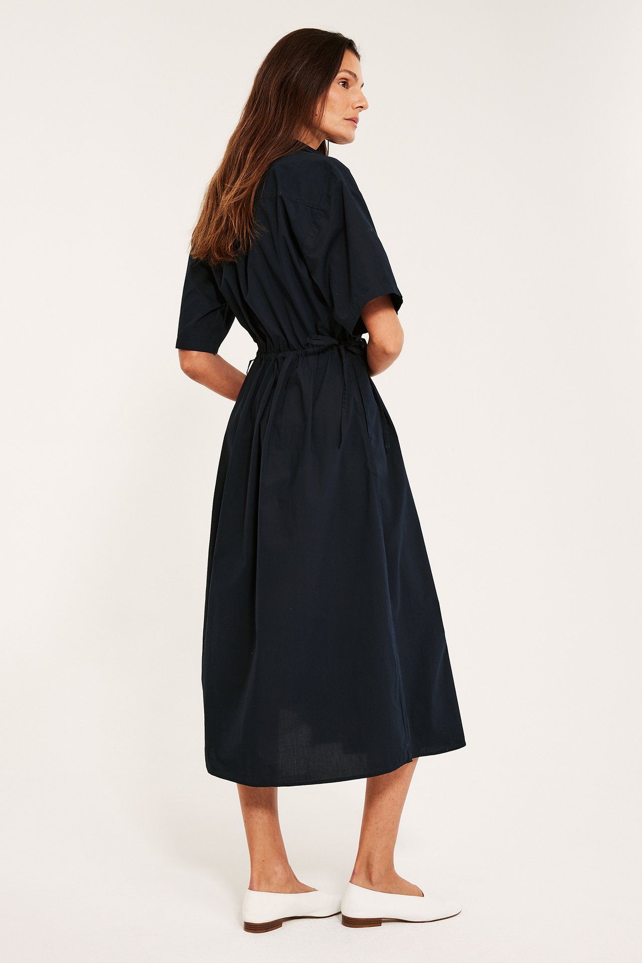 Sidovy av en kvinna i en mörkblå bomulls midiskjortklänning med dragsko i midjan och vita platta skor.