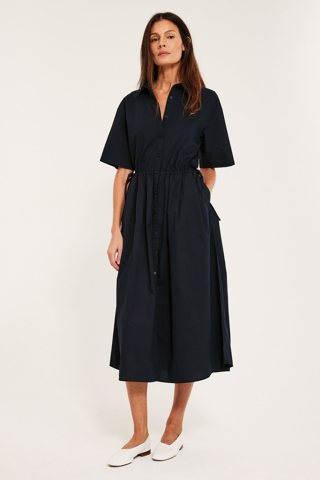 Edestäpäin, nainen tummansinisessä puuvillapaitamekossa, kiristysnyörillä, napeilla edessä ja valkoisissa kengissä.