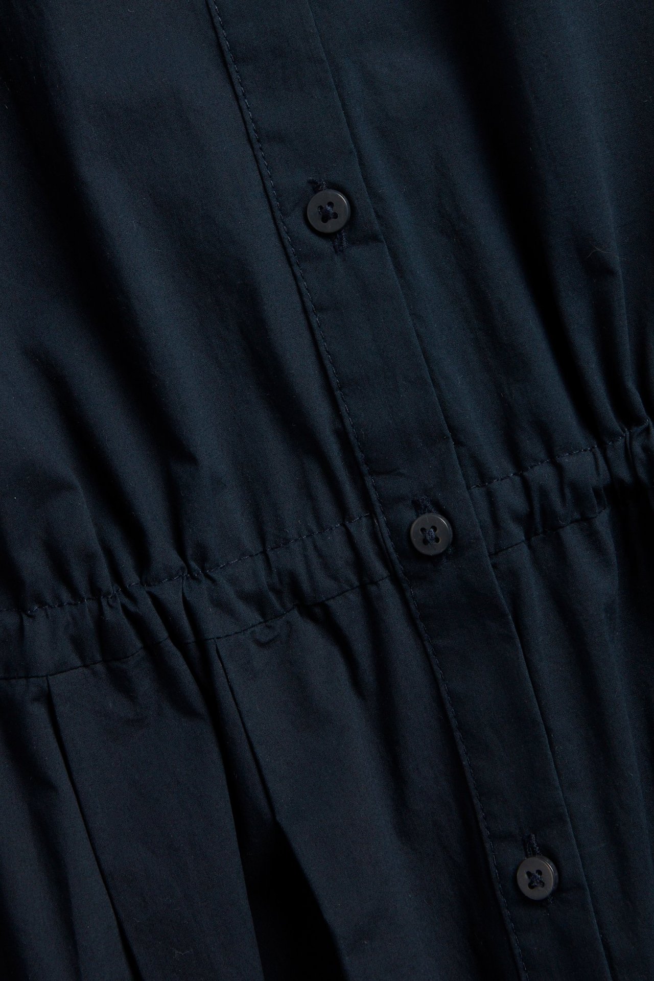 Zbliżenie ciemnoniebieskiej bawełnianej sukienki koszulowej z guzikami z przodu i widocznym ściągaczem w talii.