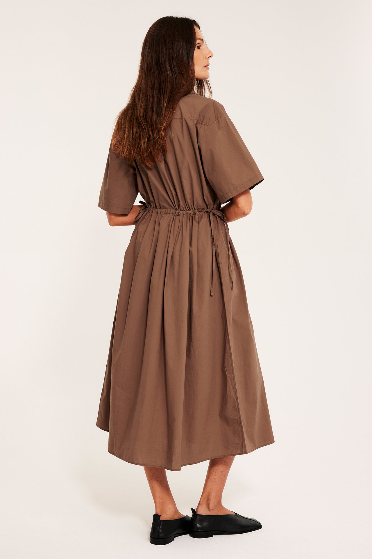 Sida-bakifrån vy: kvinna i brun bomulls midi-skjortklänning med dragsko i midjan och svarta platta skor.