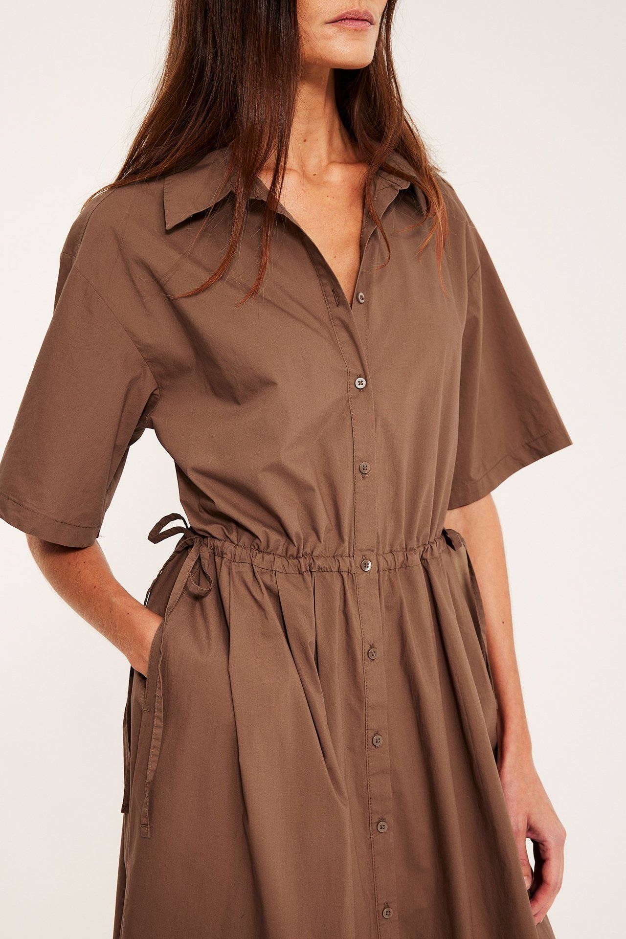 Lähikuva edestä: Nainen ruskeassa puuvillaisessa paitamekossa, lyhyet hihat, nappikiinnitys, kiristysnyöri vyötäröllä.