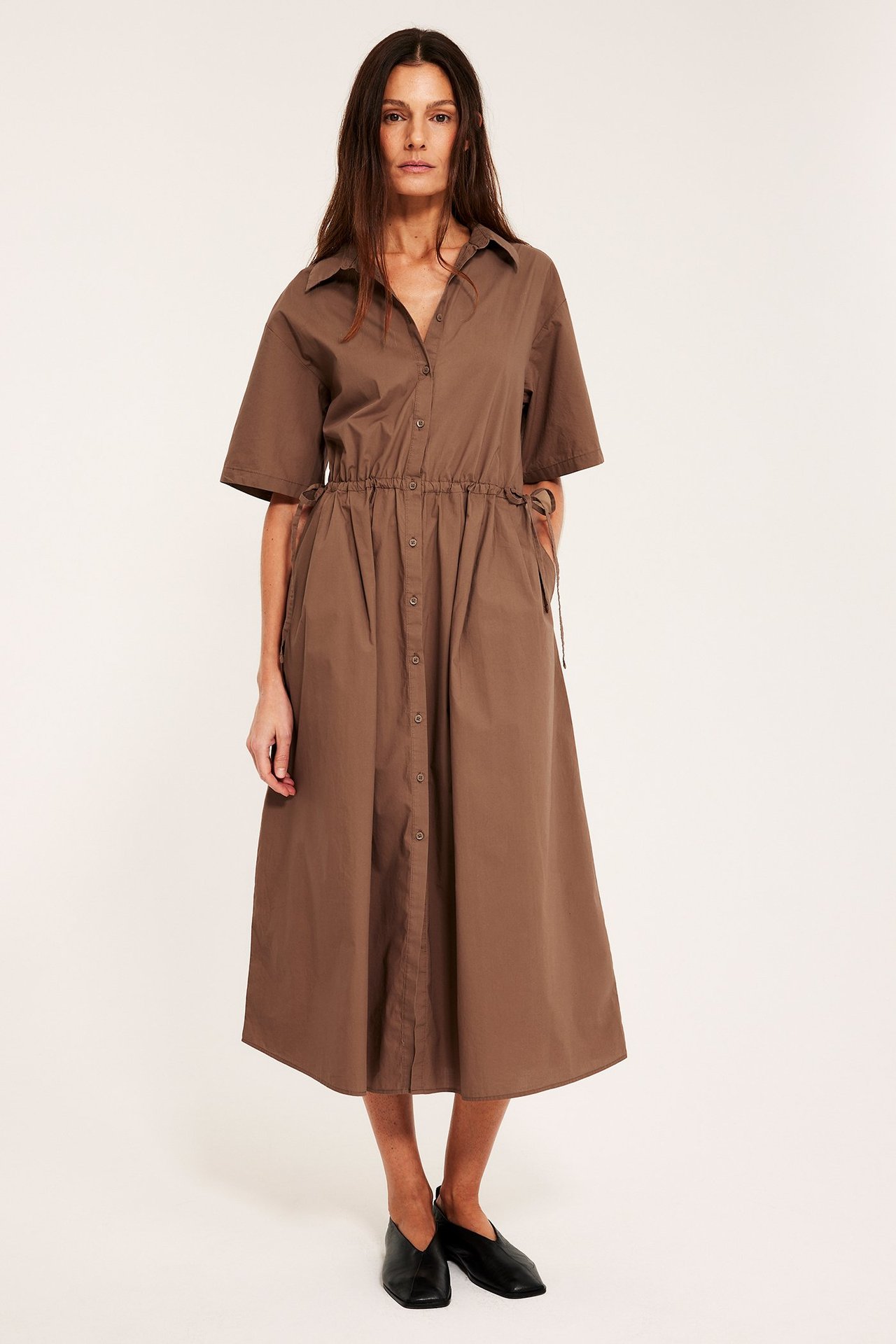 Edestä: Nainen ruskeassa puuvillapaidamekossa, kiristysnyöri vyötäröllä, napit edessä ja mustat kengät.