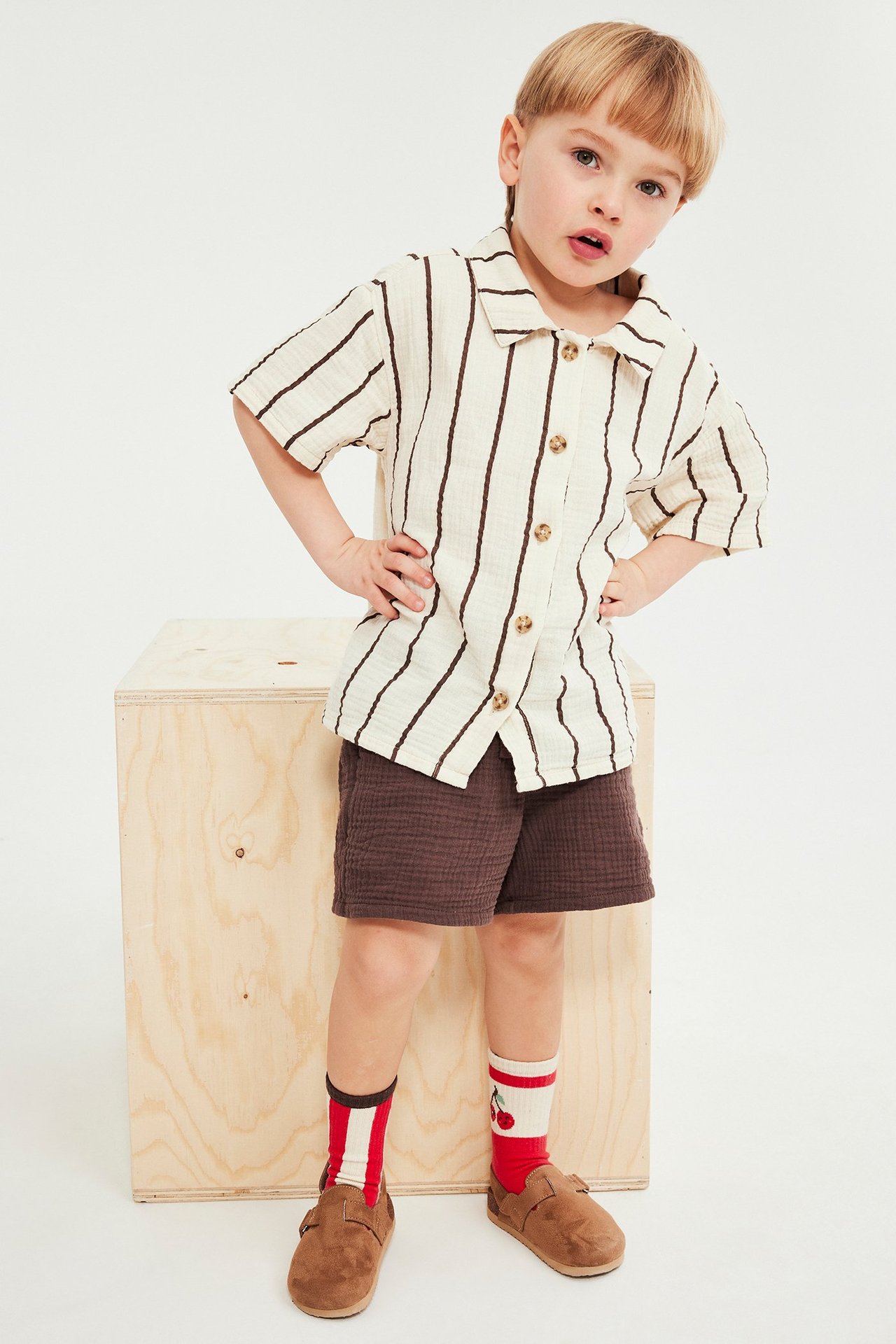 Framsida: Barn i randig musselinskjorta, bruna shorts, färgglada strumpor och bruna skor.