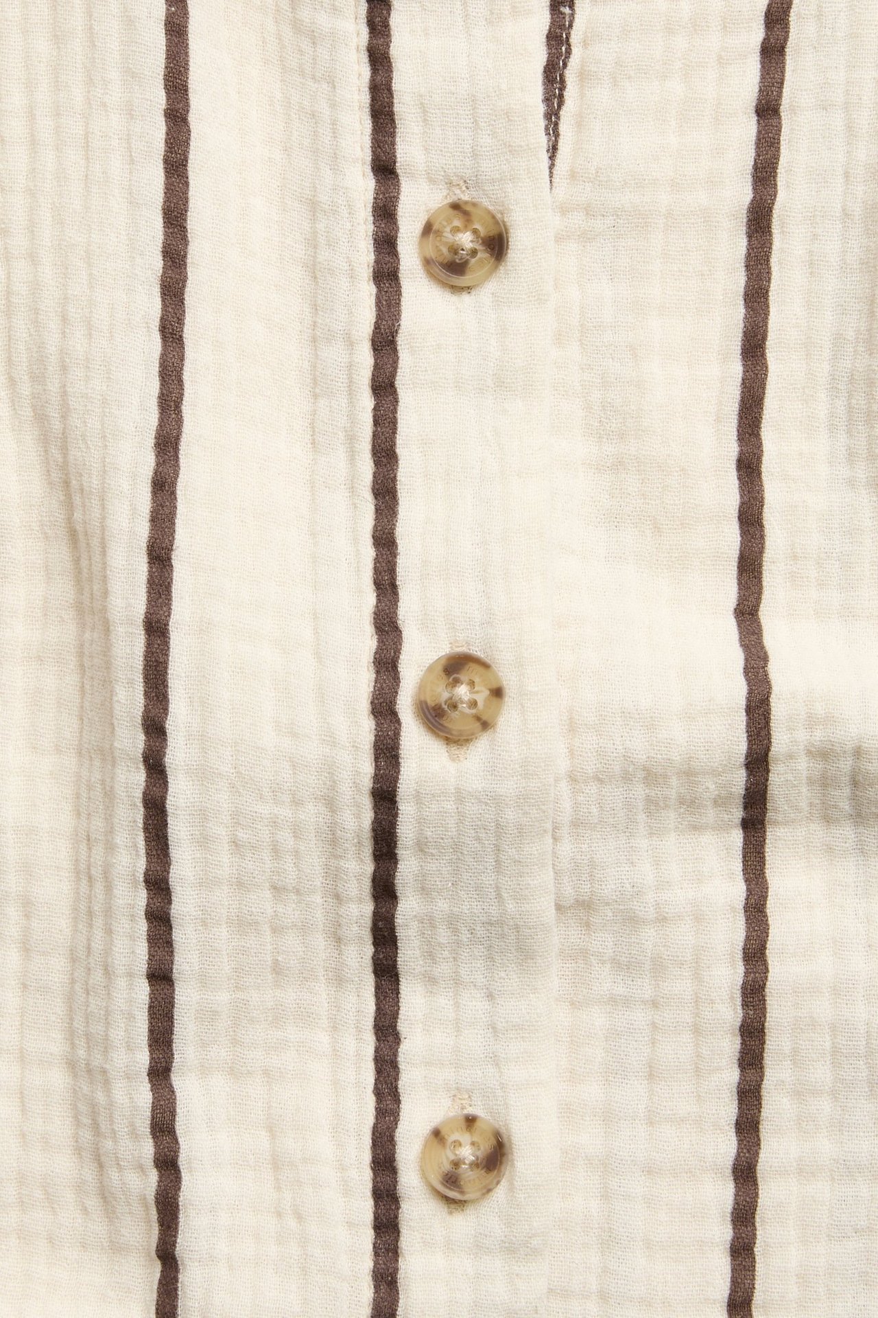 Närbild framifrån på en krämvit barnskjorta i muslin med bruna ränder och ljusbruna knappar.