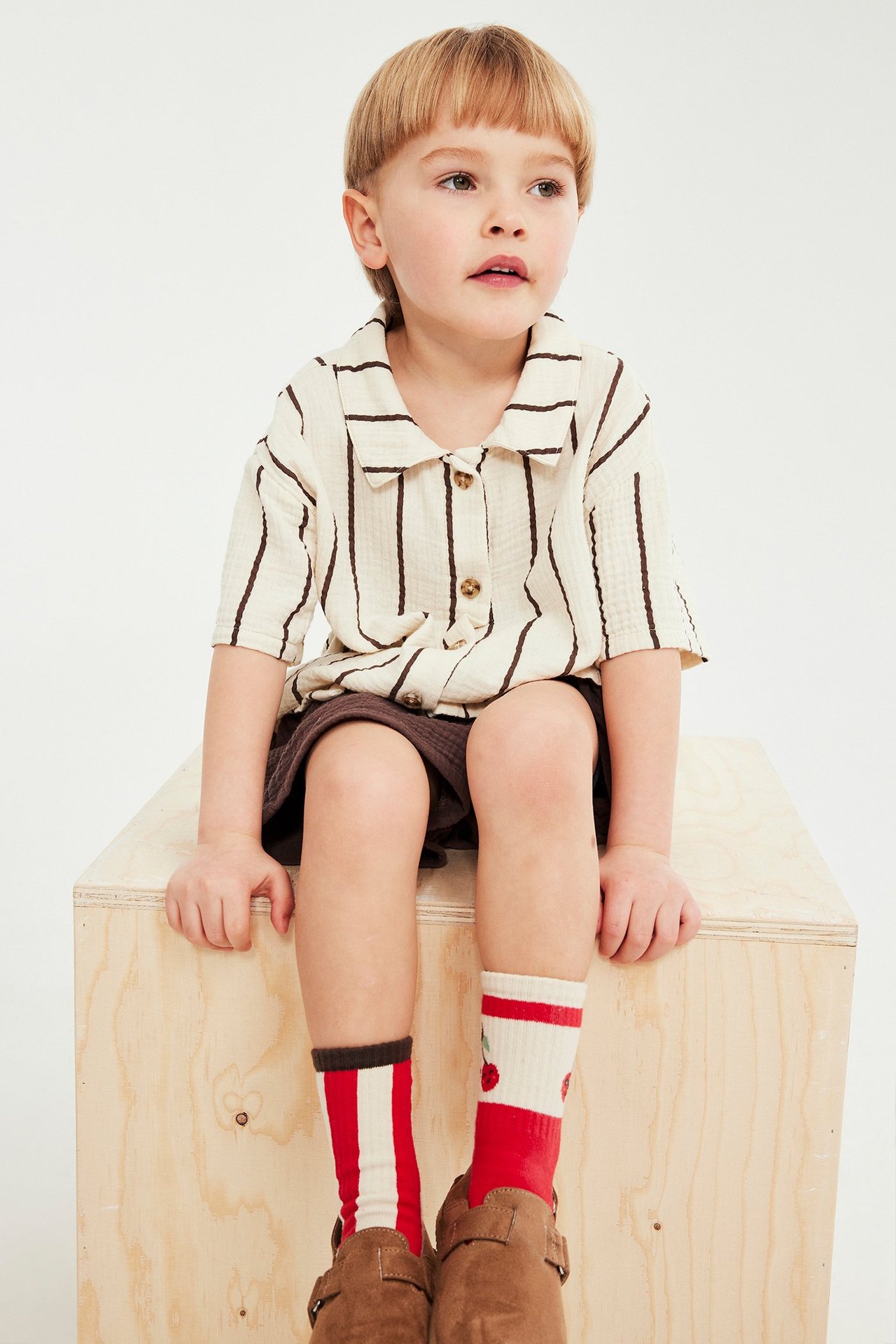 Framifrån, ett barn i brun- och beigerandig musslinskjorta, bruna shorts, omaka strumpor och bruna träskor.