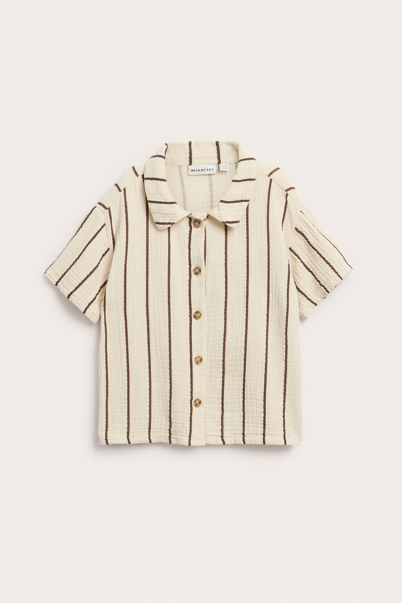 Edestä: Lasten kermanvärinen lyhythihainen musliinipaita ruskeilla pystyraidoilla ja edessä napeilla.