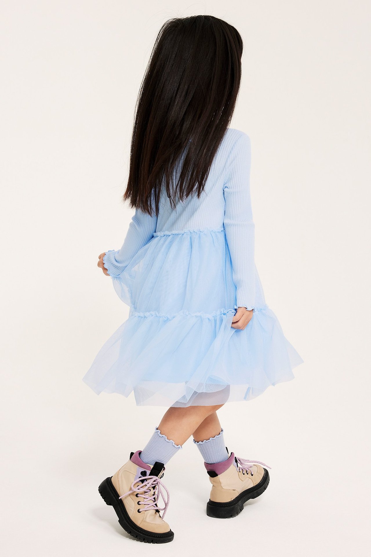 Takaapäin tyttö yllään vaaleansininen ribattu mekko tyllihameella, vaaleansiniset röyhelösukat ja kaksiväriset saappaat.