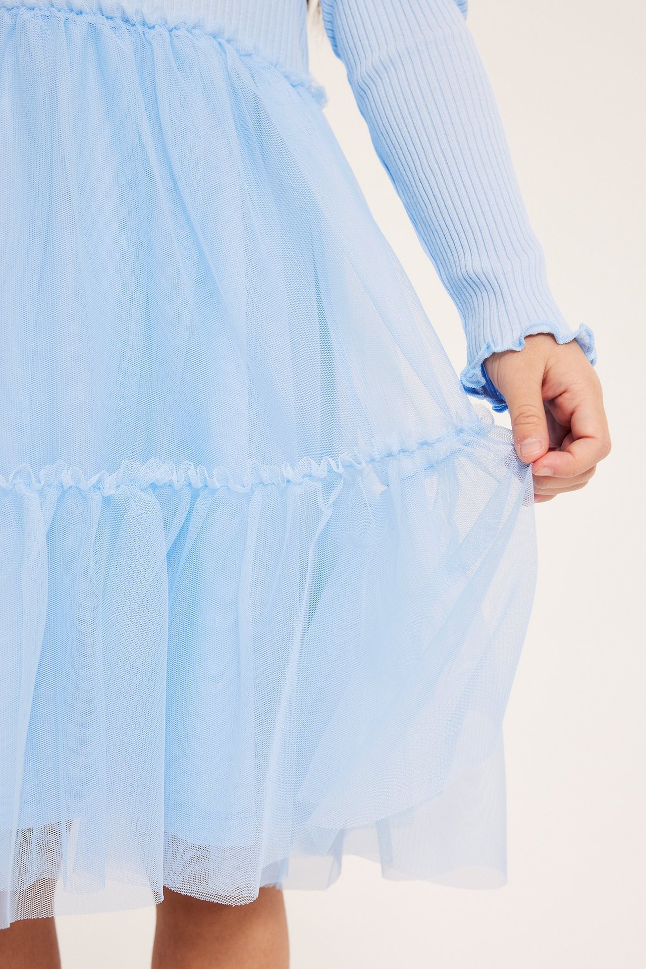 Vaaleansininen lasten mekko, jossa ribbineulottu yläosa ja kerrostettu tyllihame. Käsi pitelee helmaa.