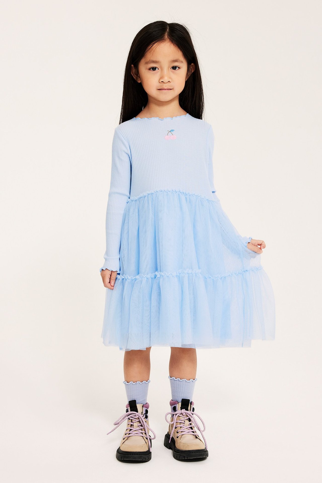Widok z przodu: dziecko w jasnoniebieskiej sukience z prążkowaną górą, tiulową spódnicą, haftem, skarpetkami i beżowymi butami.