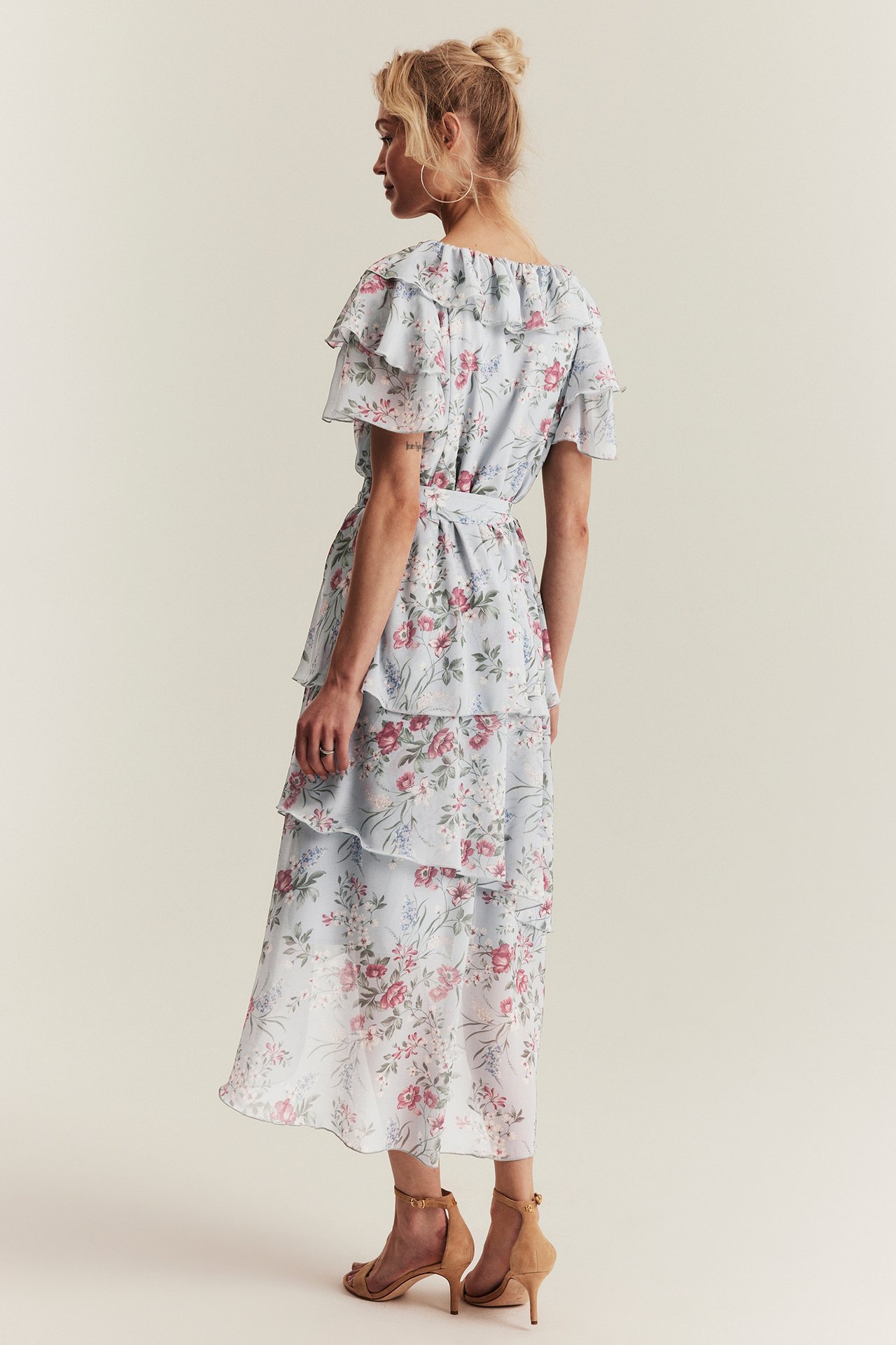 Widok z tyłu kobiety w jasnoniebieskiej kwiecistej szyfonowej sukience z falbanami, z beżowymi szpilkami.