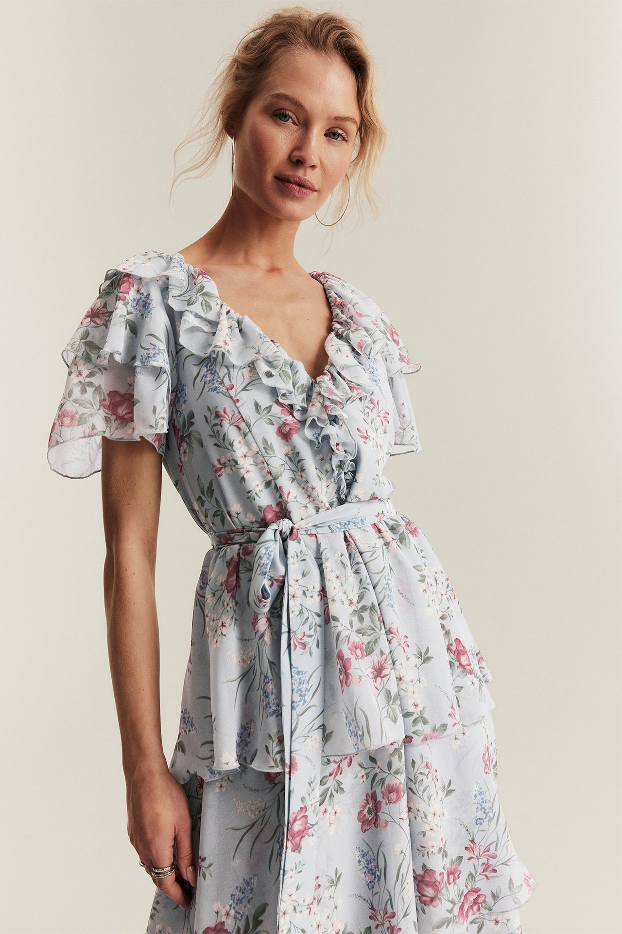 Frontvy av kvinna i ljusblå blommig chiffongklänning med volanger, korta ärmar och knytskärp.