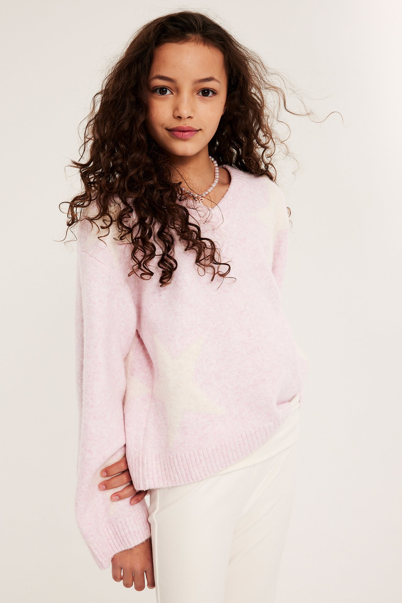 Frontal: Jente iført lys rosa v-hals strikket genser med hvite stjerner og hvite bukser.