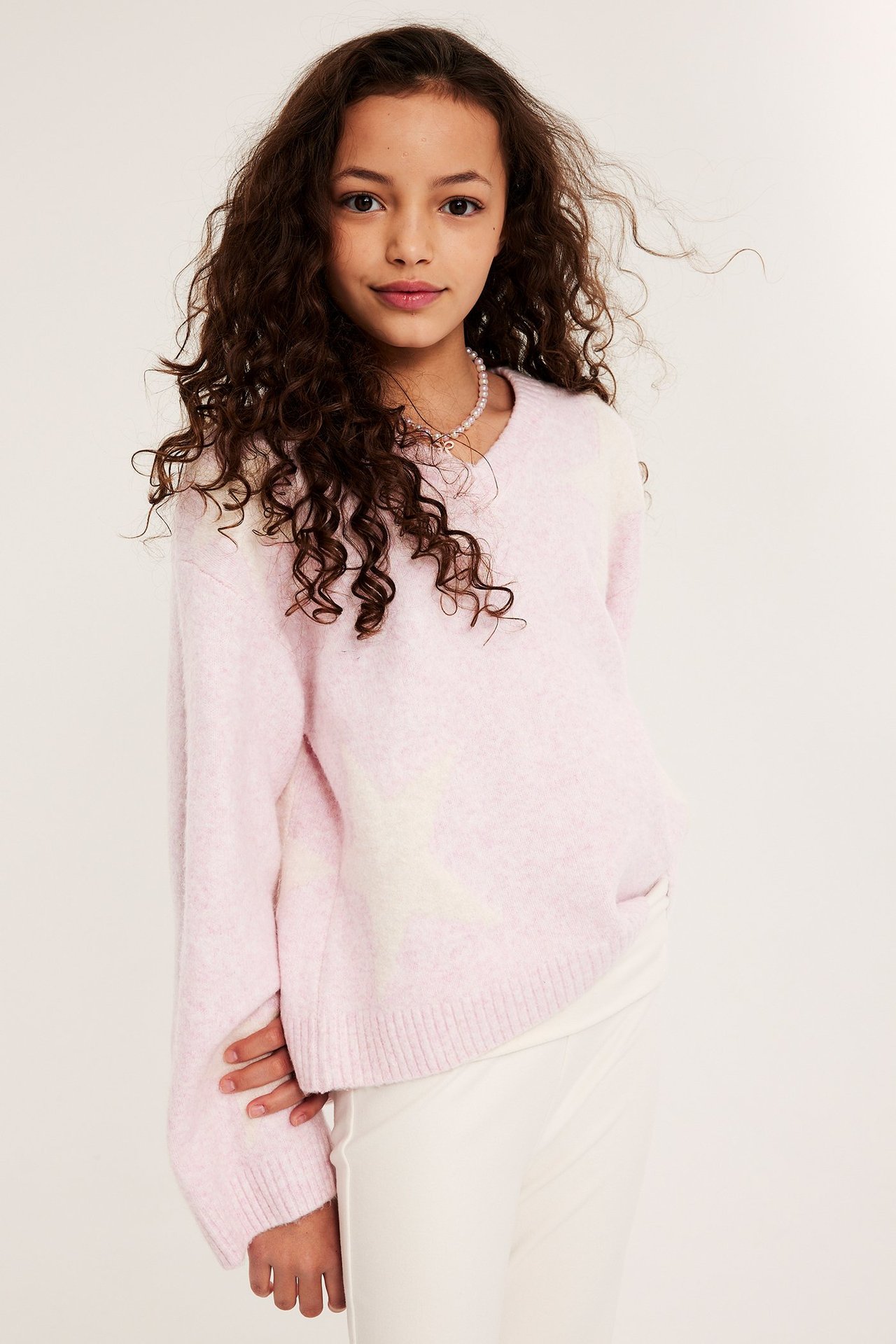 Edestä: Tyttölapsi vaaleanpunaisessa V-kaula-aukkoisessa neuleessa valkoisilla tähdillä ja valkoisissa housuissa.