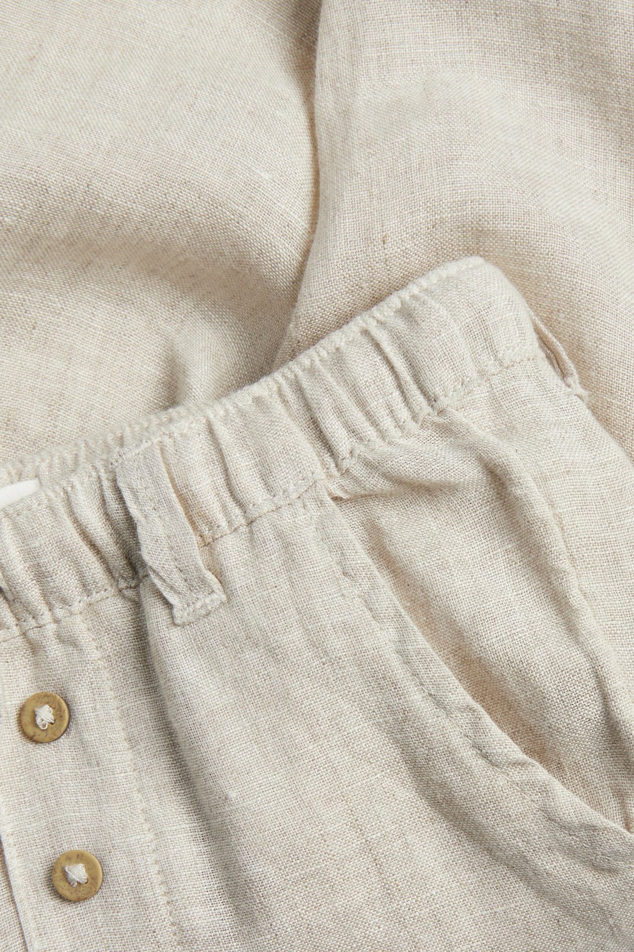 Närbild av ljusbeige linnebyxor med elastisk midja och dekorativ knappgylf.