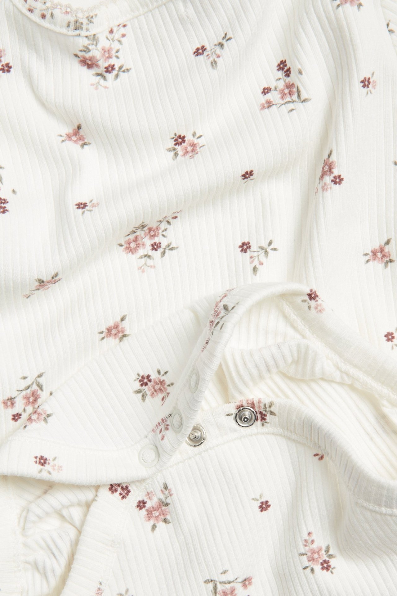Lähikuva vauvan luonnonvalkoisesta ribbineulebodysta, jossa kukkakuvio ja painonapit olkapäällä.