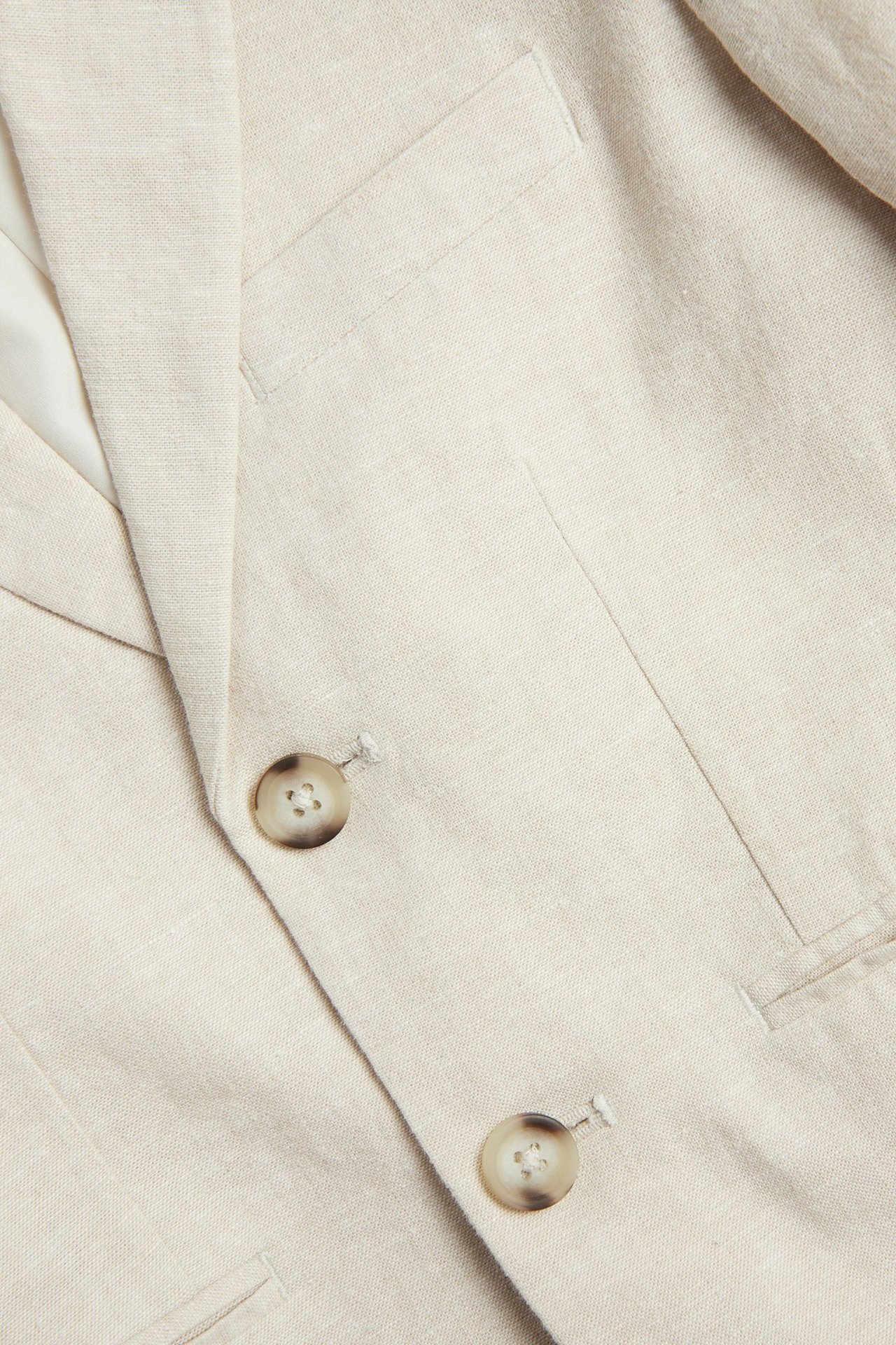 Zbliżenie na jasnobeżową marynarkę z domieszką lnu, z dwoma guzikami i kieszeniami.