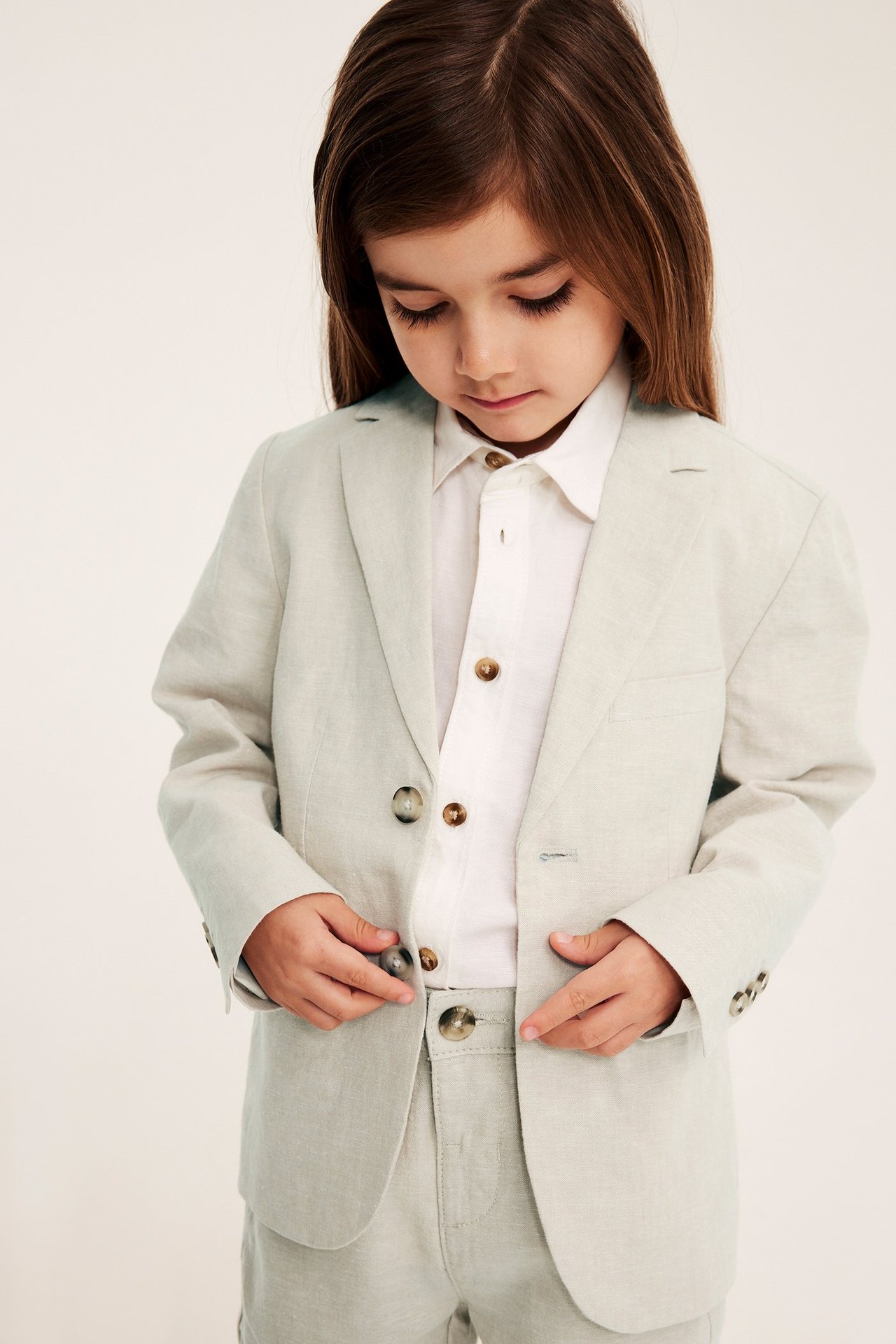 Forfra: Barn i lys beige linblazer, hvit skjorte og matchende bukse, hender på knapper.