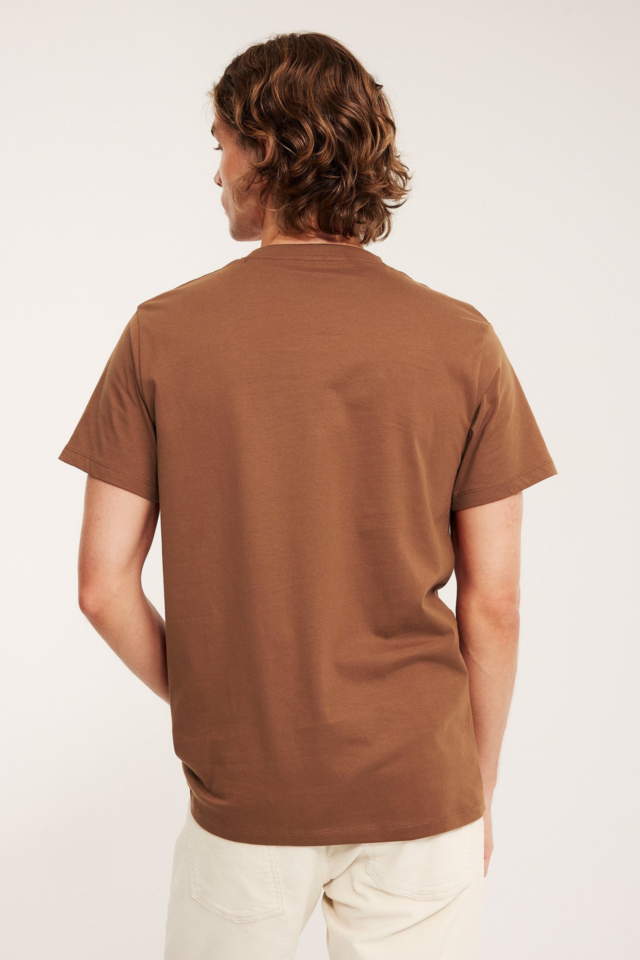 Bakfra sett, mann i brun, ensfarget rundhalset t-skjorte og lysebeige bukser.