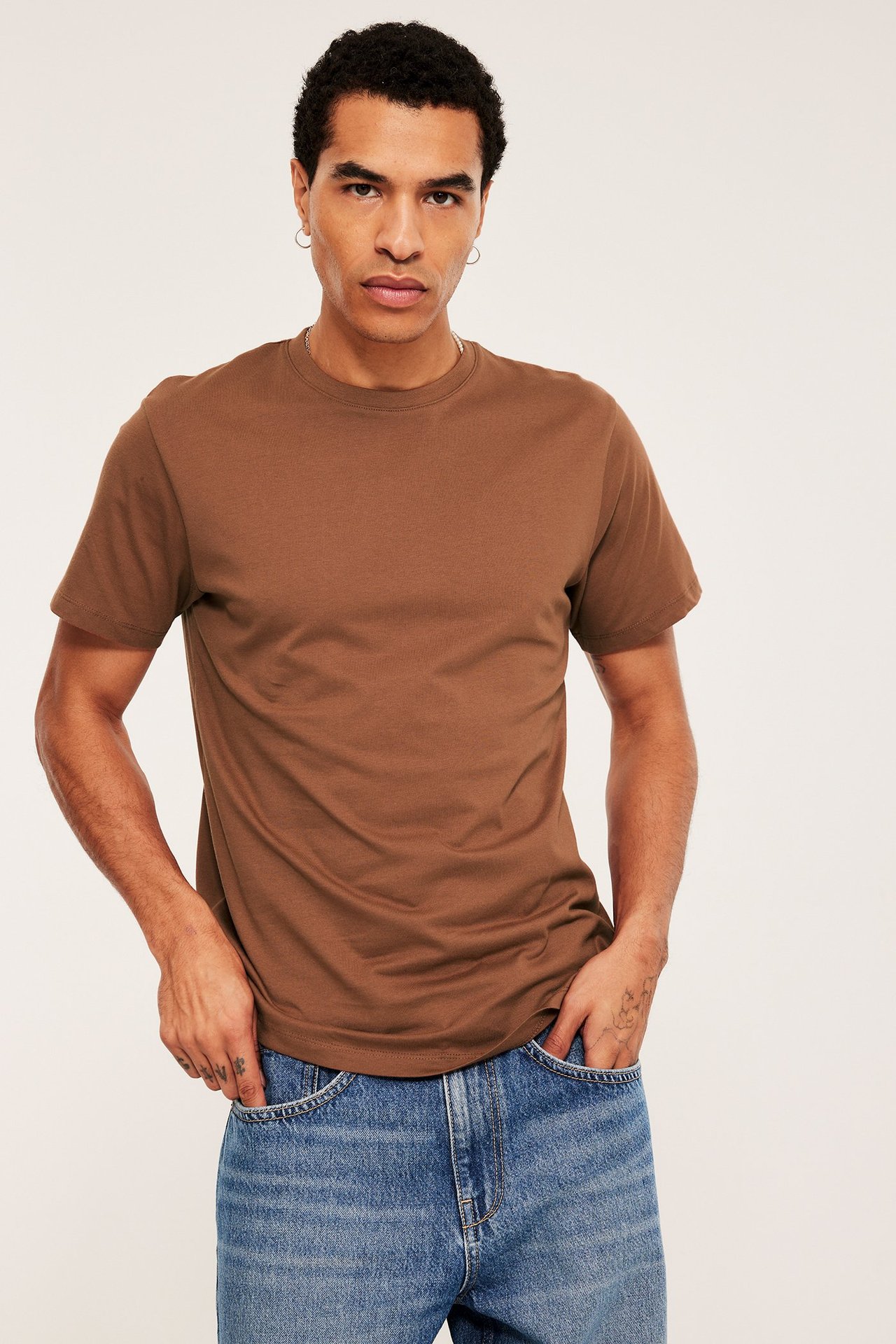 Edestäpäin, mies yllään ruskea yksivärinen pyöreäkauluksinen puuvilla-t-paita ja siniset farkut kädet taskuissa.