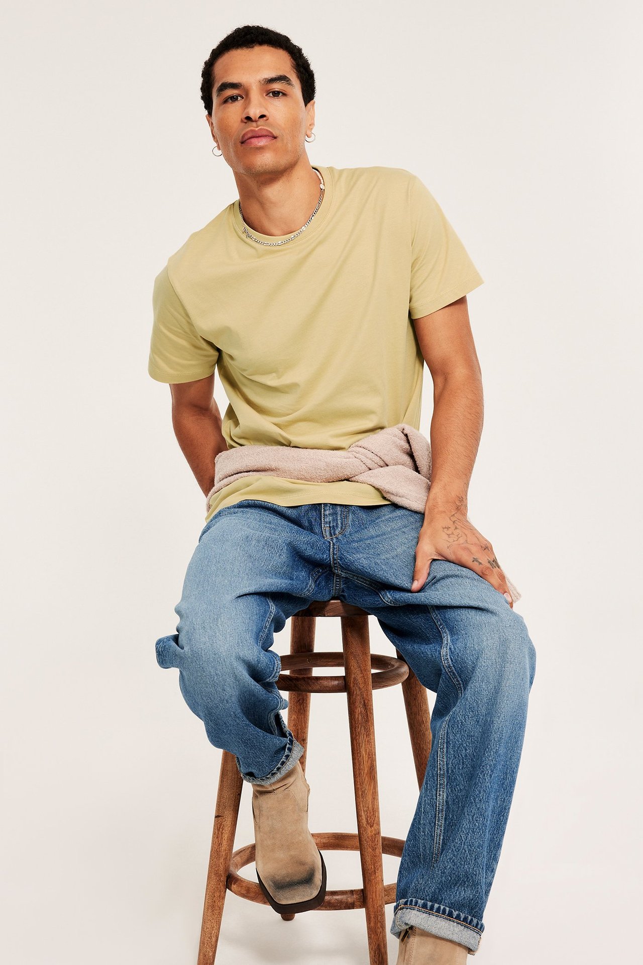 Mann i lys gul t-skjorte, blå jeans, beige genser i livet, lysebrune støvler.