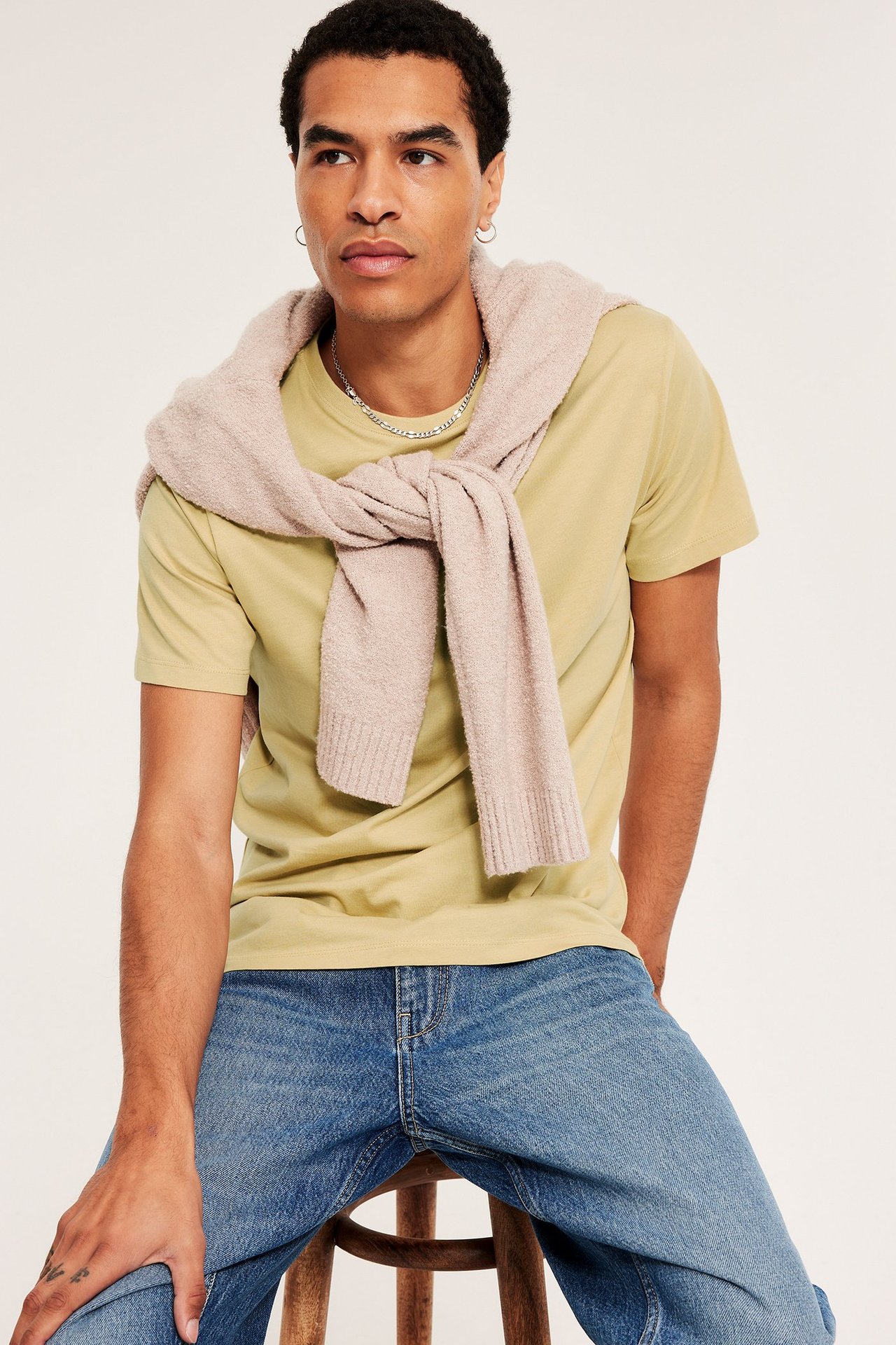 Edestä: Mies vaaleankeltaisessa puuvilla-t-paidassa, sinisissä farkuissa ja beigessä neuleessa olkapäillä.