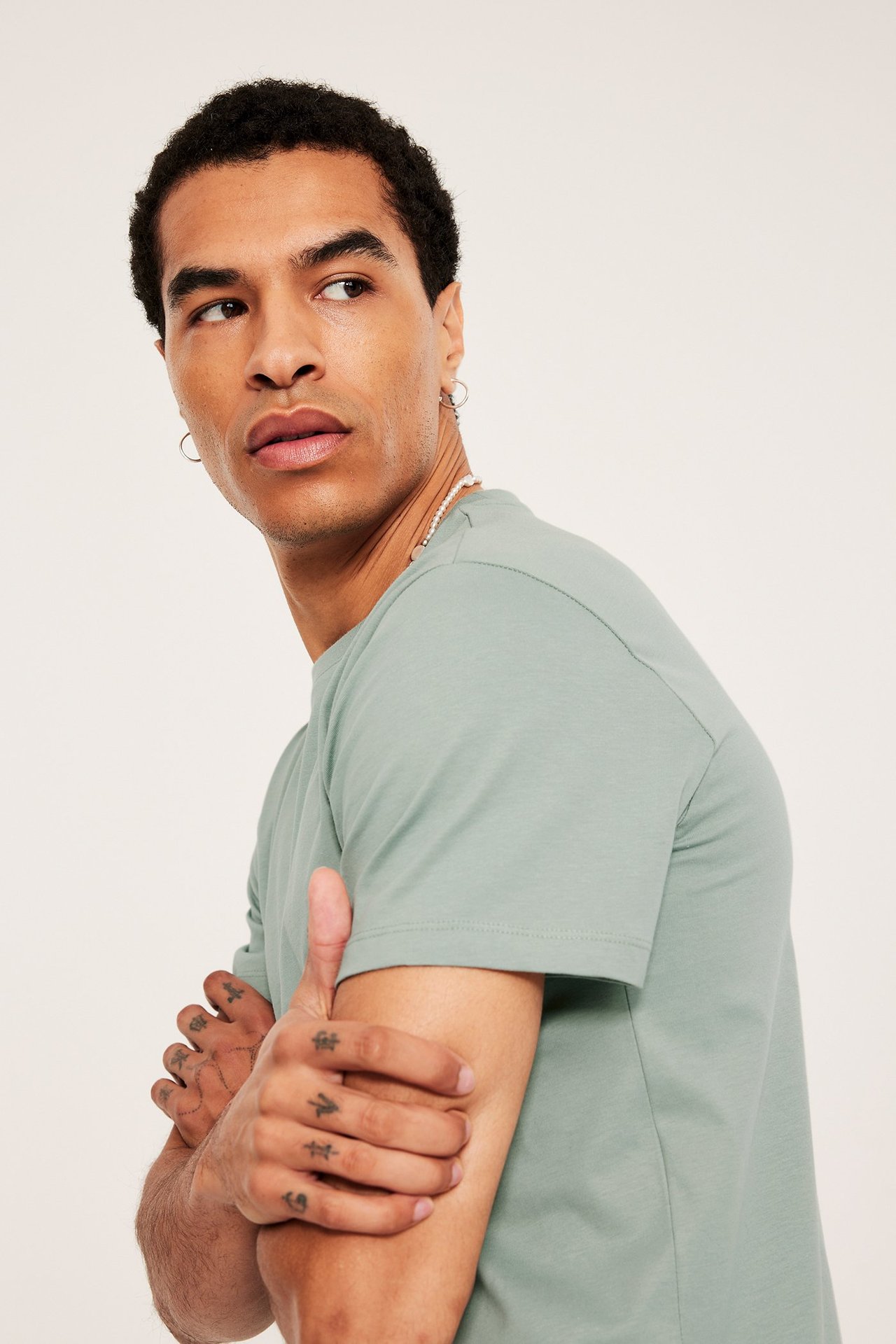 Nærbilde sideprofil av mann i lysegrønn ensfarget rundhalset T-skjorte, sølvringer i ørene og perlekjede.
