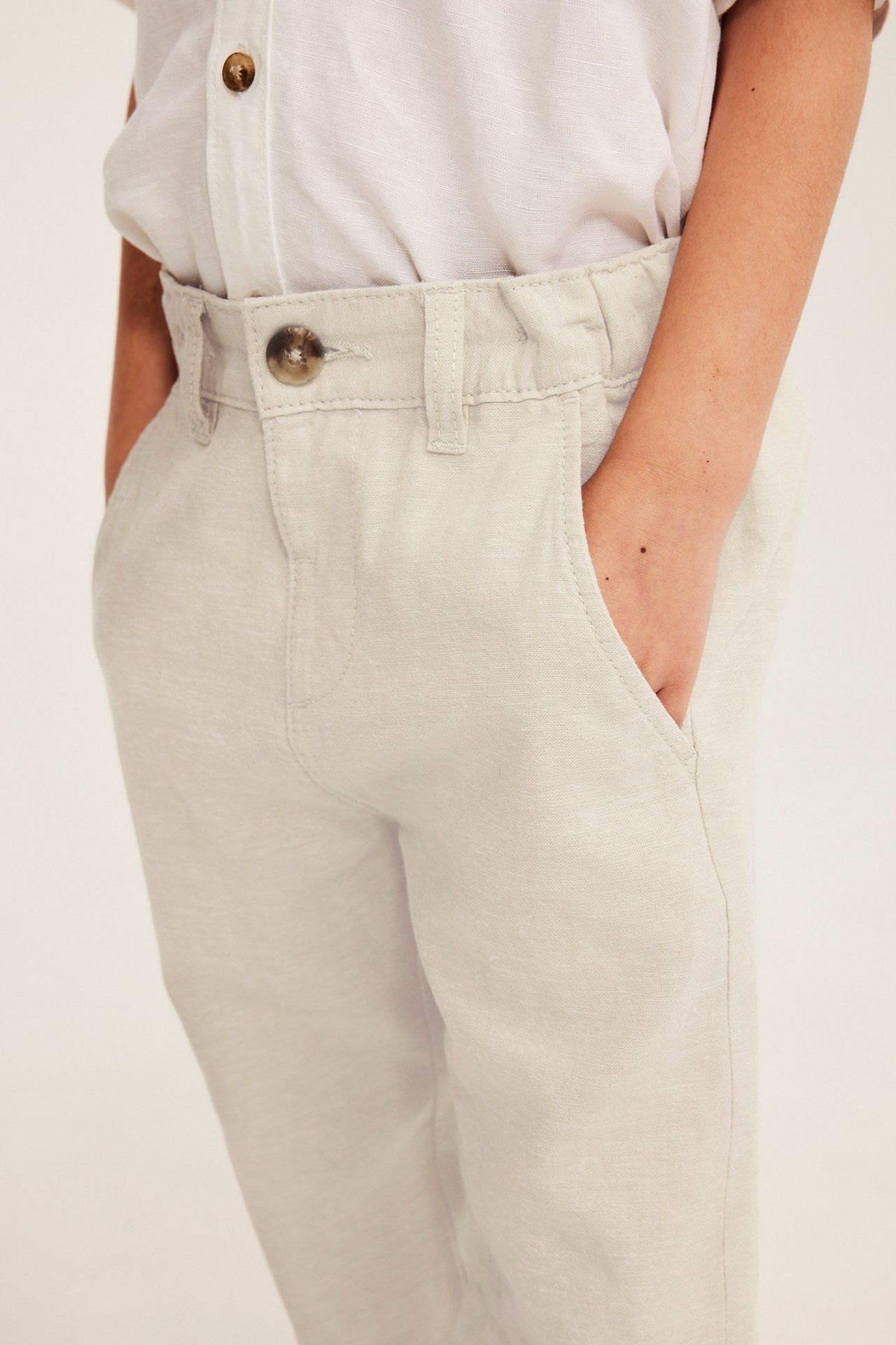 Frontvy: Barn i ljusbeige linnebyxor med knapp, hand i fickan och en ljus skjorta.