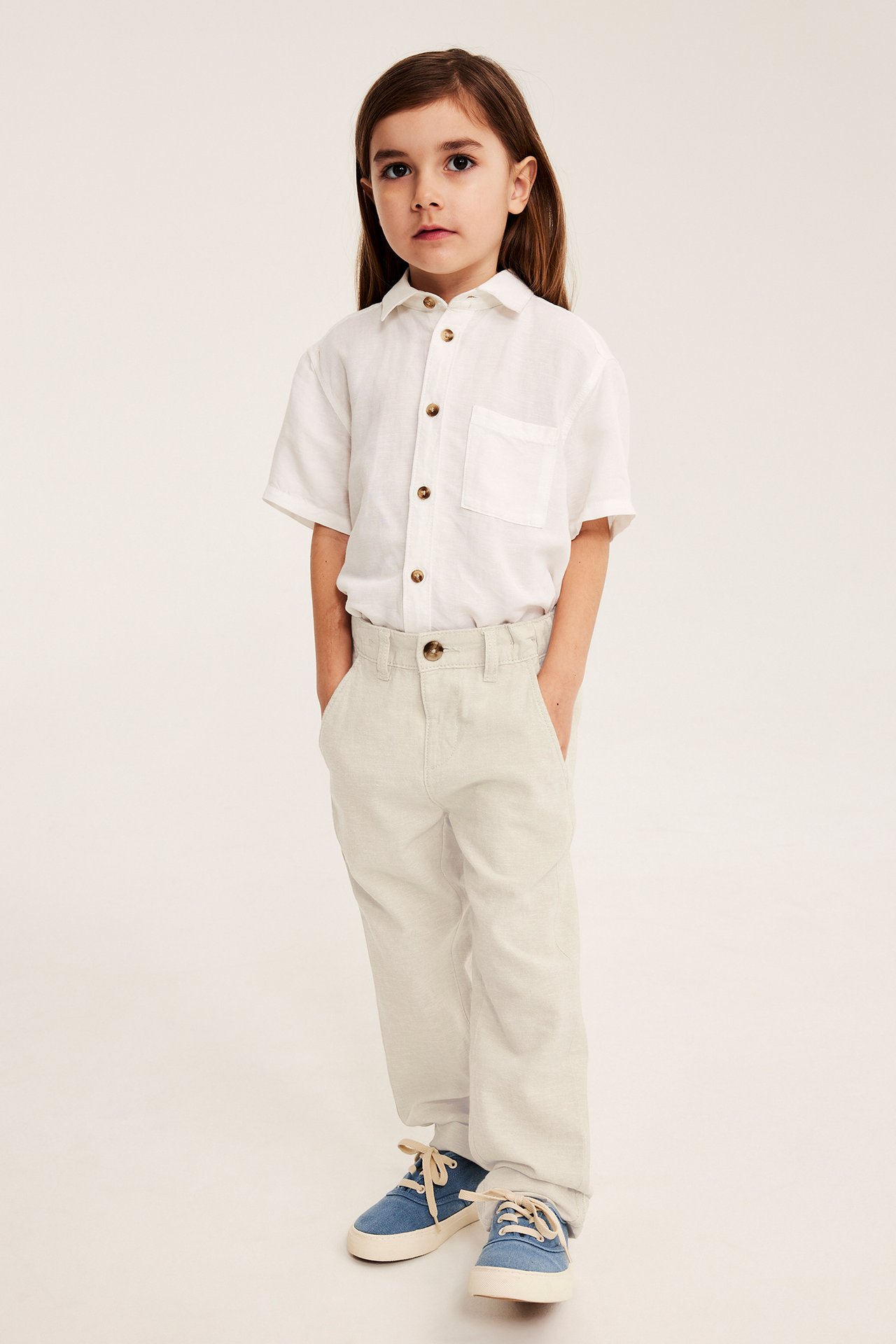 Framsida: Kid bär ljusbeige linneblandade byxor med fickor, knapp, vit kortärmad skjorta och blå sneakers.