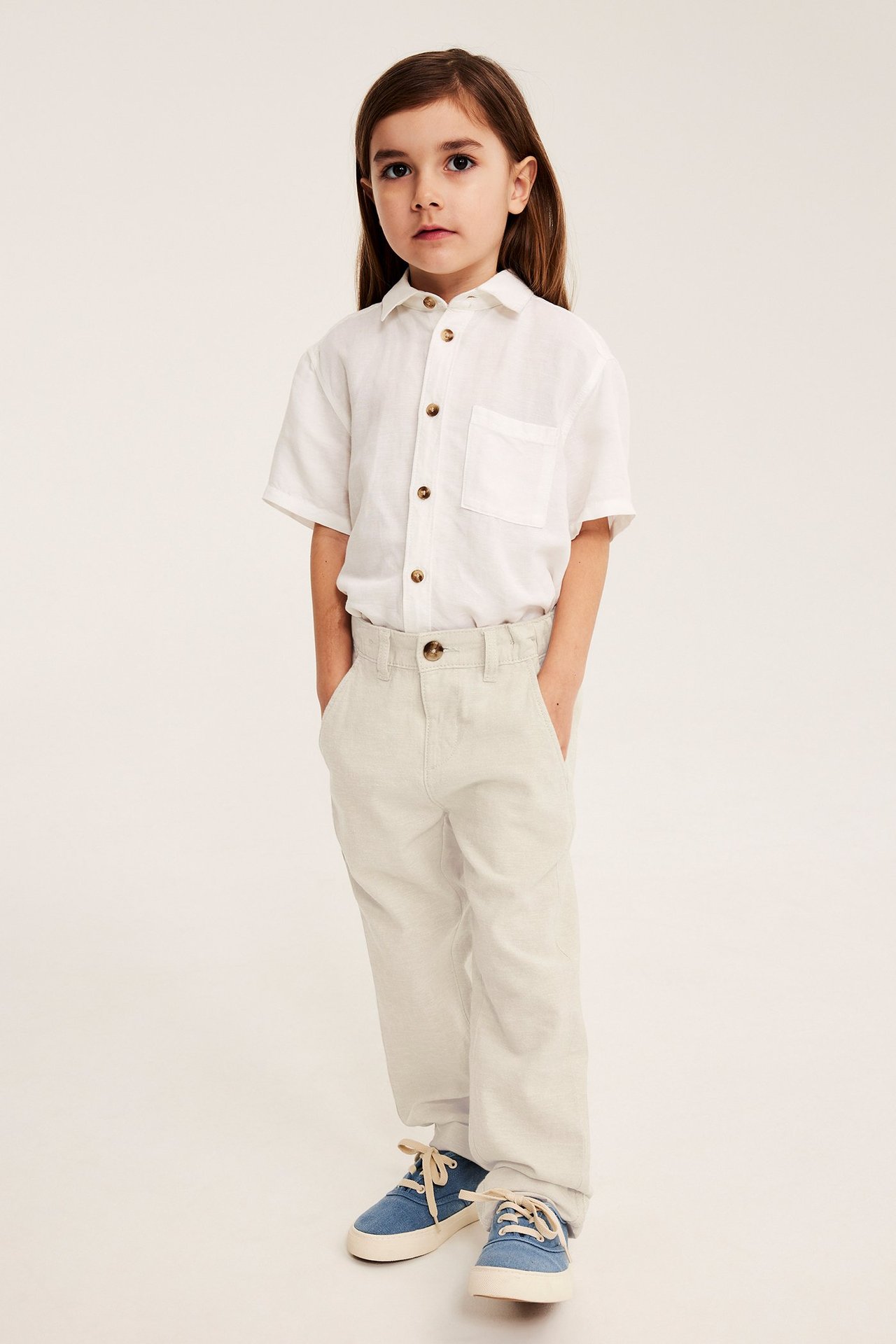 Widok z przodu: Dziecko w jasnobeżowych spodniach z mieszanki lnu, białej koszuli i niebieskich trampkach.