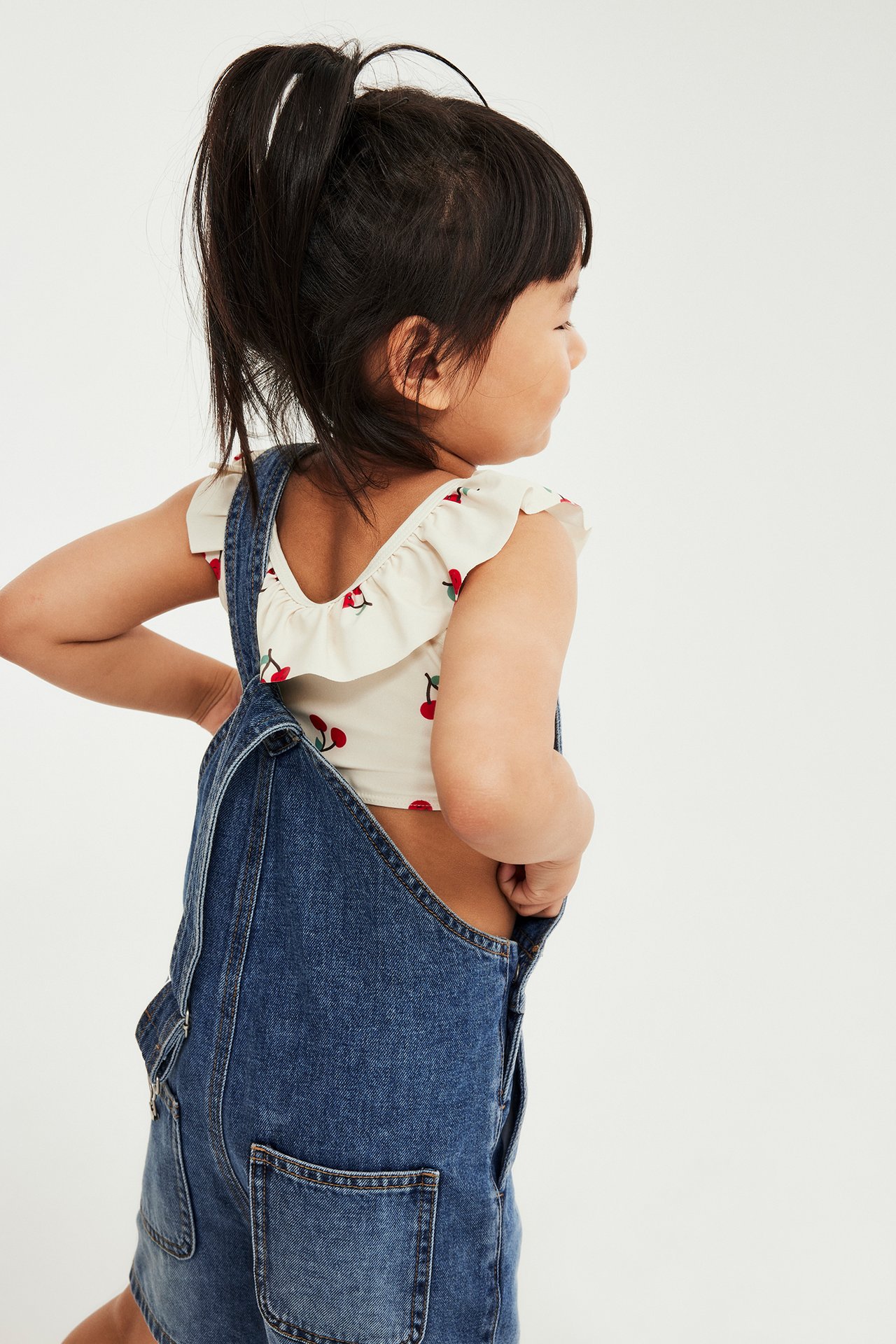 Ryggvy: Barn i ljusbeige körsbärsmönstrad volangbikiniöverdel under blå denimoveraller.