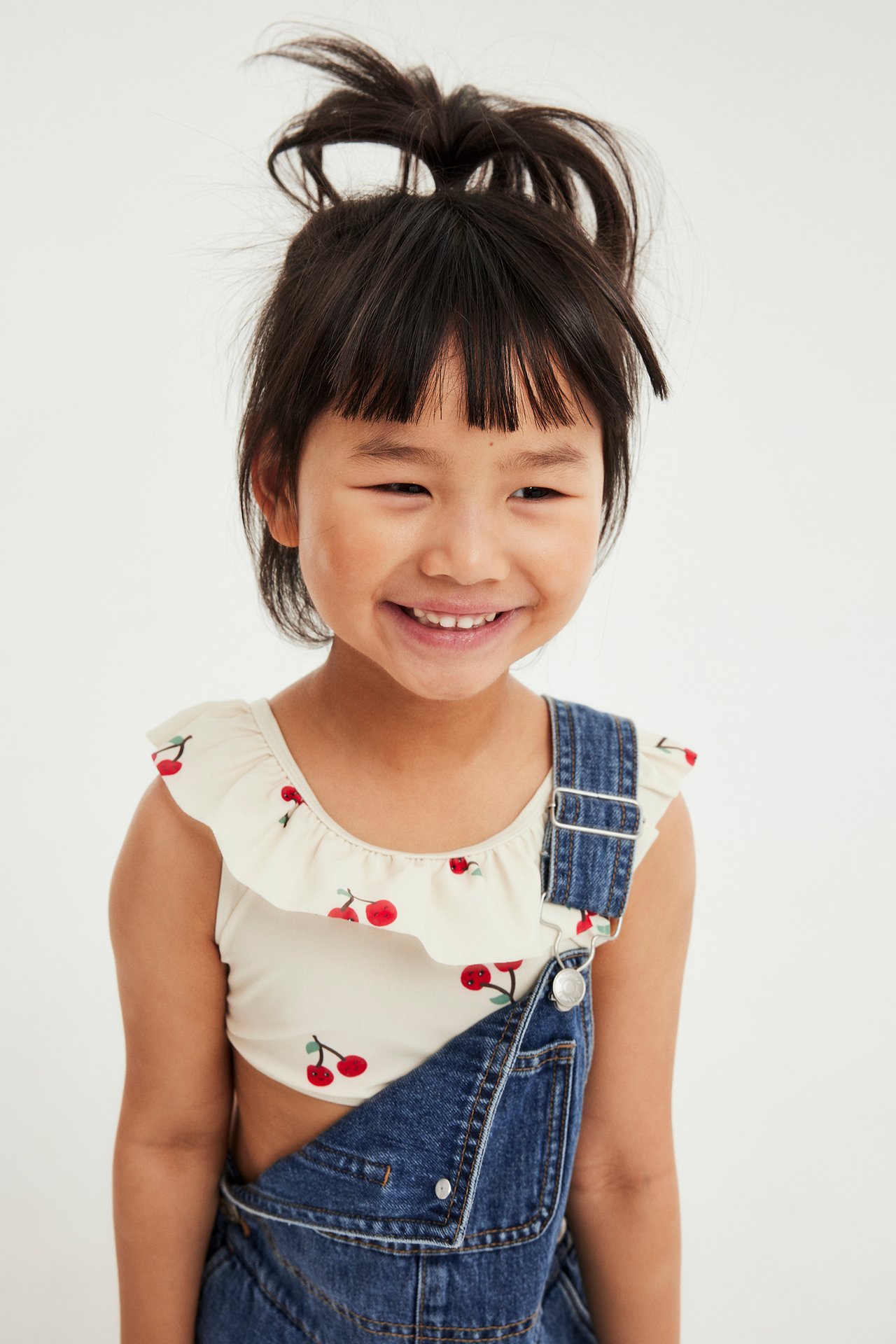 Framifrån, ett leende barn i ljusbeige volangbikinitopp med körsbärsmönster under blå denimoveraller.