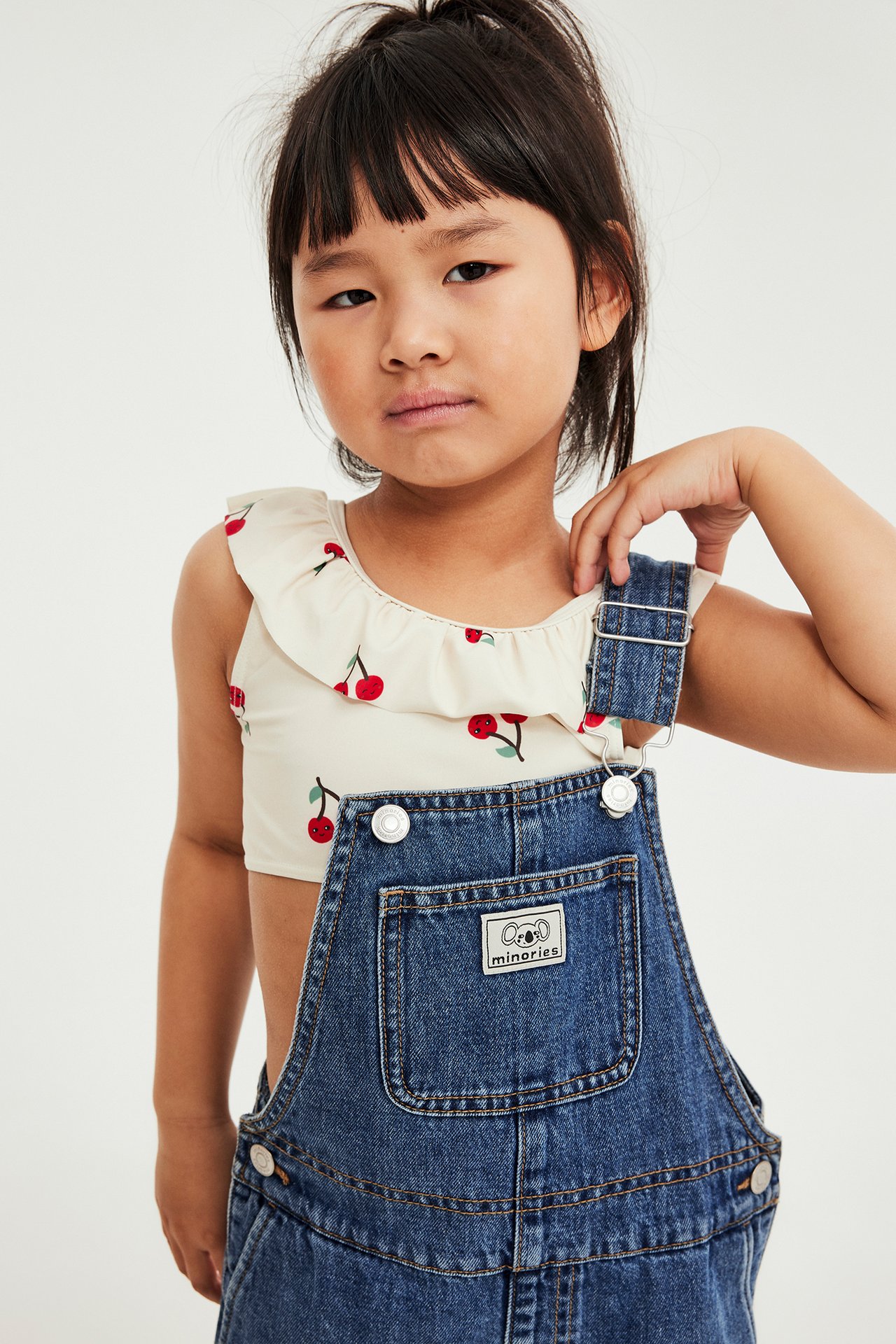 Widok z przodu dziecka w jasnobeżowym kostiumie kąpielowym z falbanką i wiśniami pod niebieskimi ogrodniczkami.