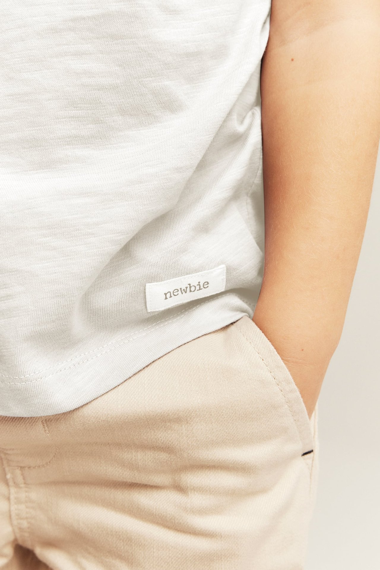 Lähikuva lapsesta off-white slub-trikoopaidassa ja vaaleanbeigessä housuissa käsi taskussa.