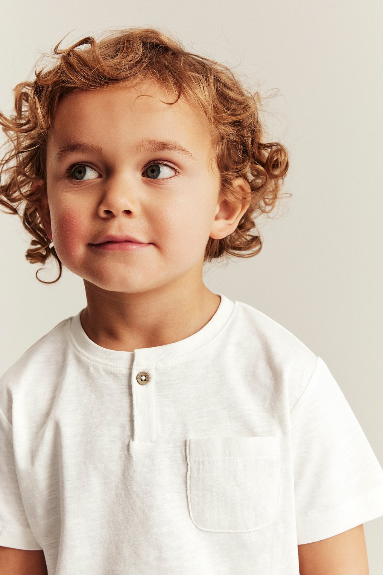 Nærbilde av et barn i en offwhite slub-jersey T-skjorte med en knapp og brystlomme.