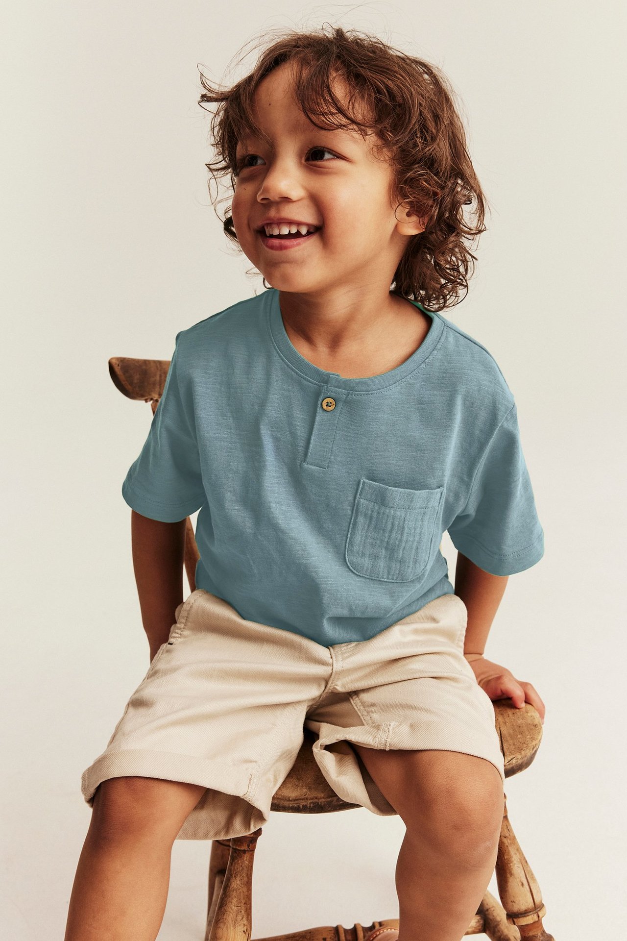 Framifrån-sidvy: Ett leende barn bär en mörkturkos bomulls-t-shirt med knapp, ficka och beige shorts.