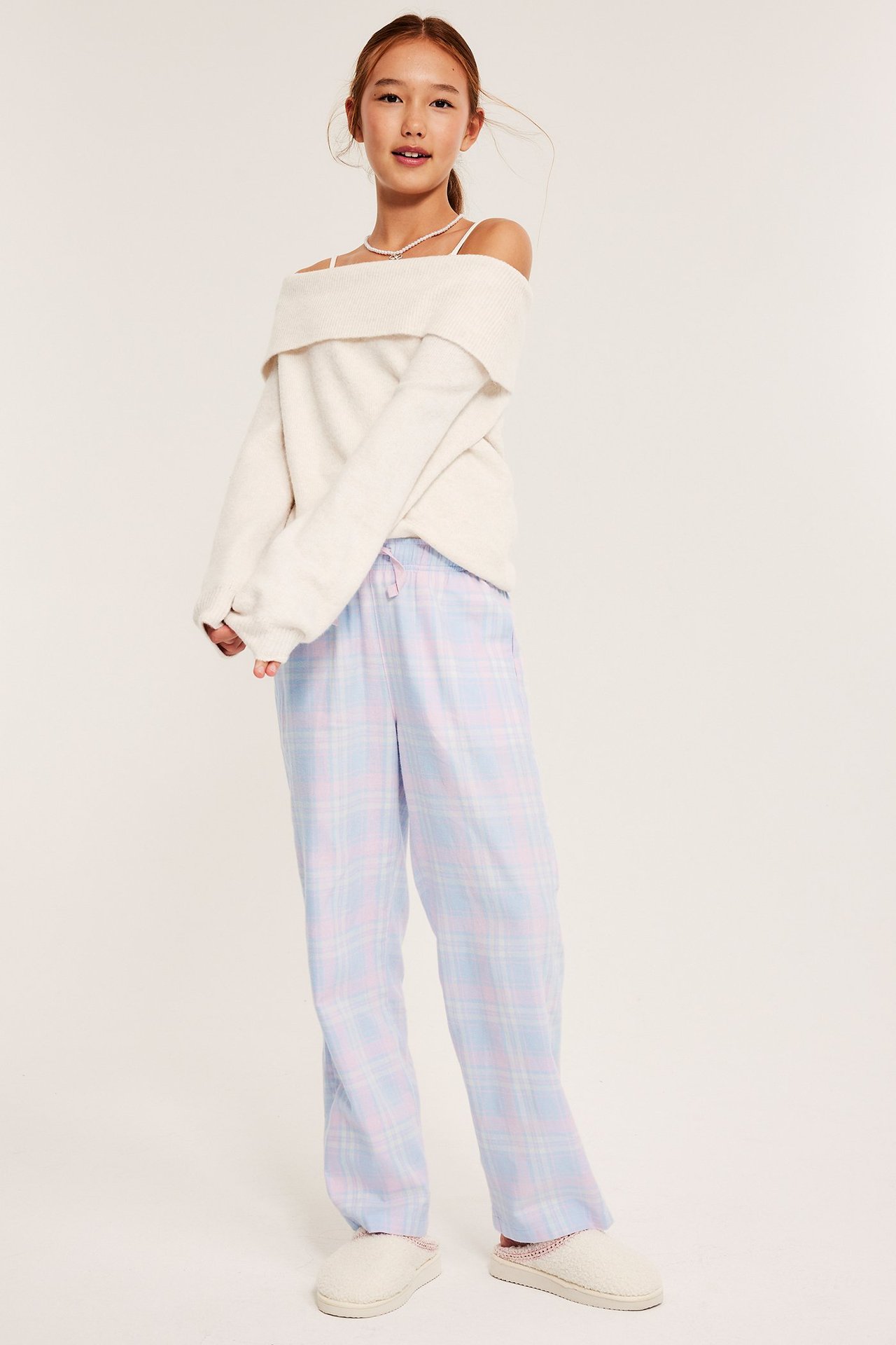 Front view av ett barn i blårutiga pyjamasbyxor i flanell, en benvit off-shoulder tröja och gräddfärgade tofflor.