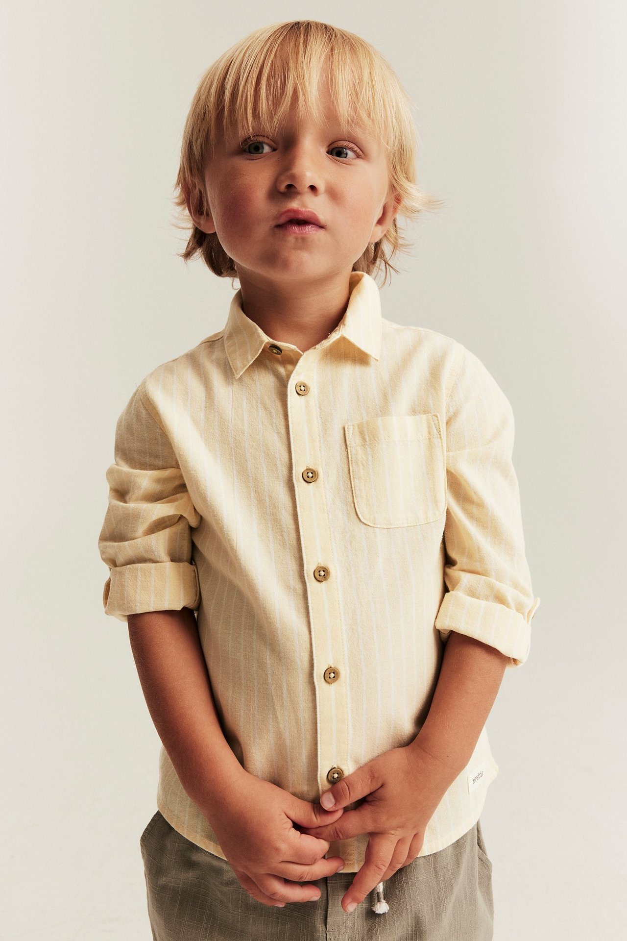 Frontvy av ett barn i en gulrandig vävd bomullsskjorta med krage, upprullade ärmar och grå byxor.