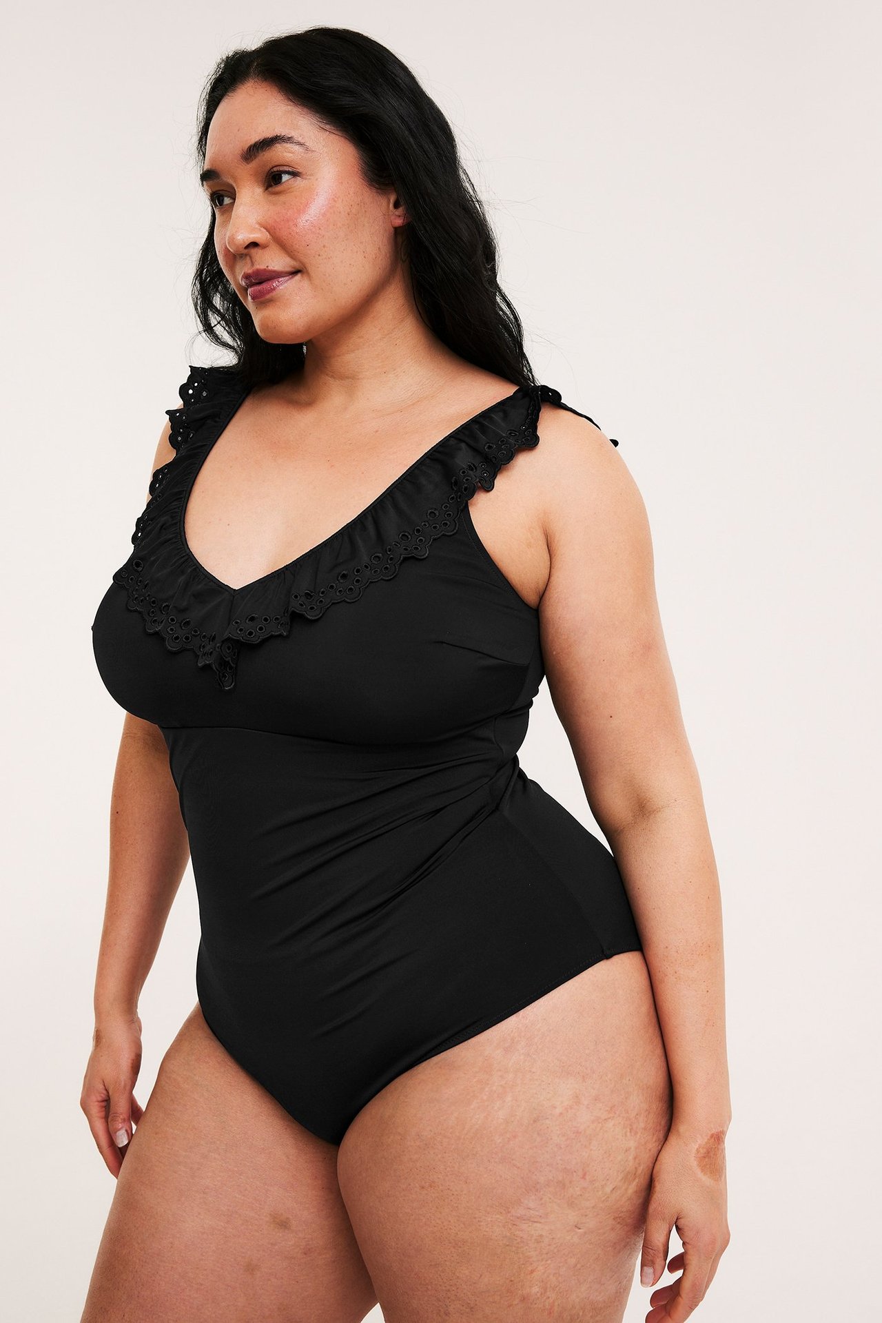 Nainen, edestäpäin, mustassa uimapuvussa, jossa reikäkoristellut röyhelöt V-pääntiessä ja olkaimissa.