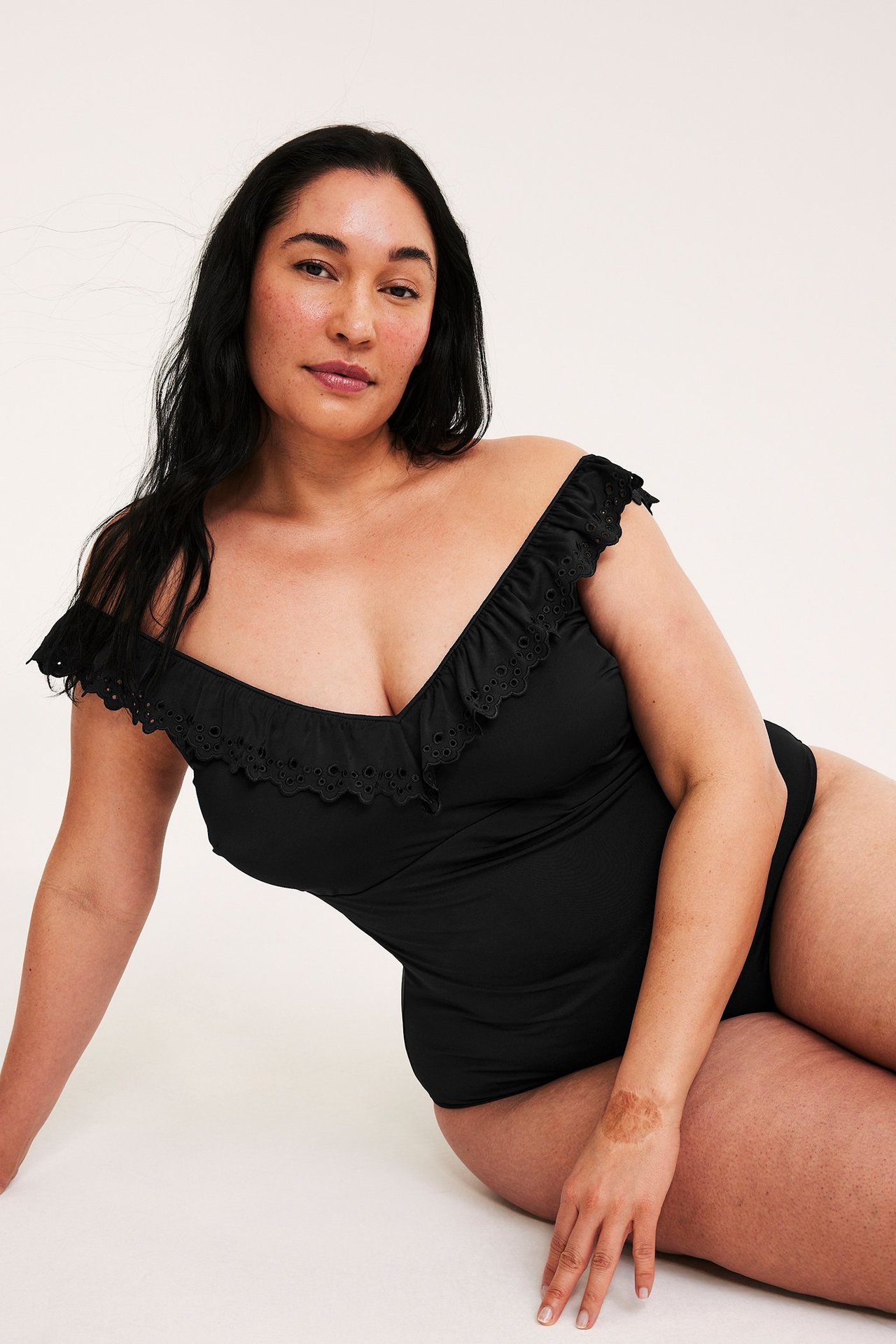 Edestä, istuva nainen mustassa uimapuvussa, jossa olkapäät paljastavat röyhelöt ja reikäkoristeet.