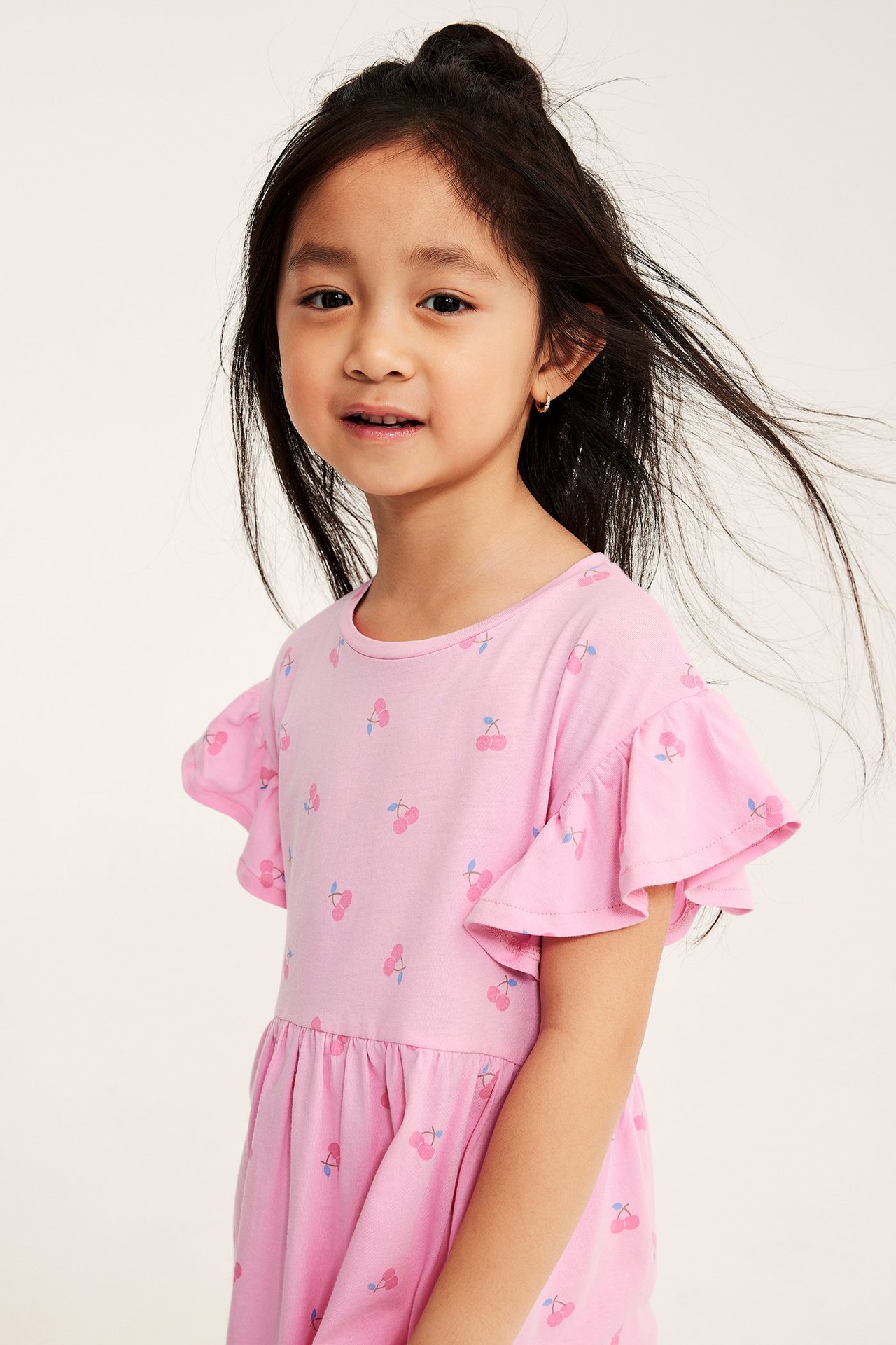 Lähikuva edestä lapsesta vaaleanpunaisessa puuvillamekossa kirsikkakuviolla ja lyhyillä röyhelöhihoilla.