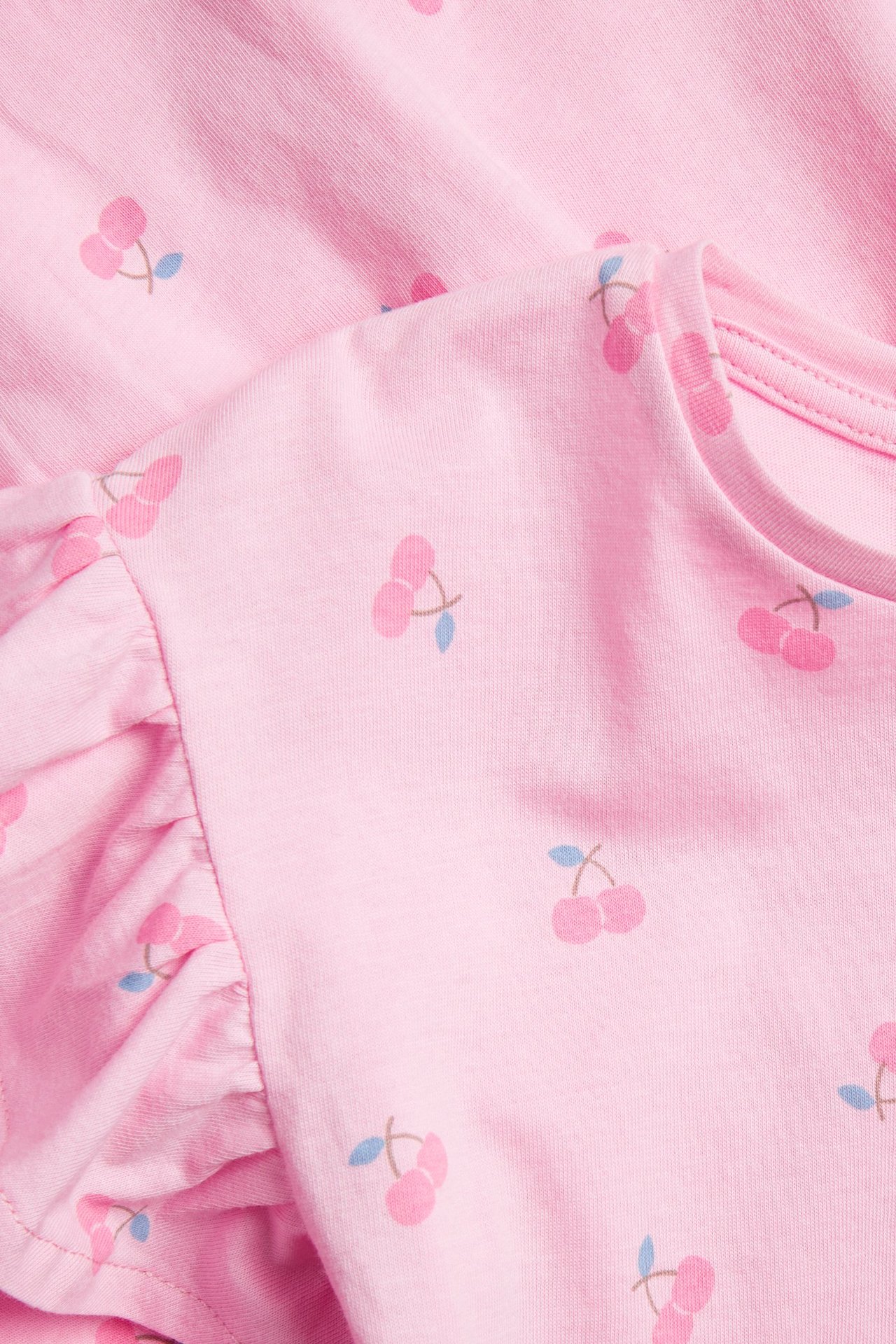 Nærbilde av en lys rosa bomullskjole for barn, med kirsebærmønster og volangerme.