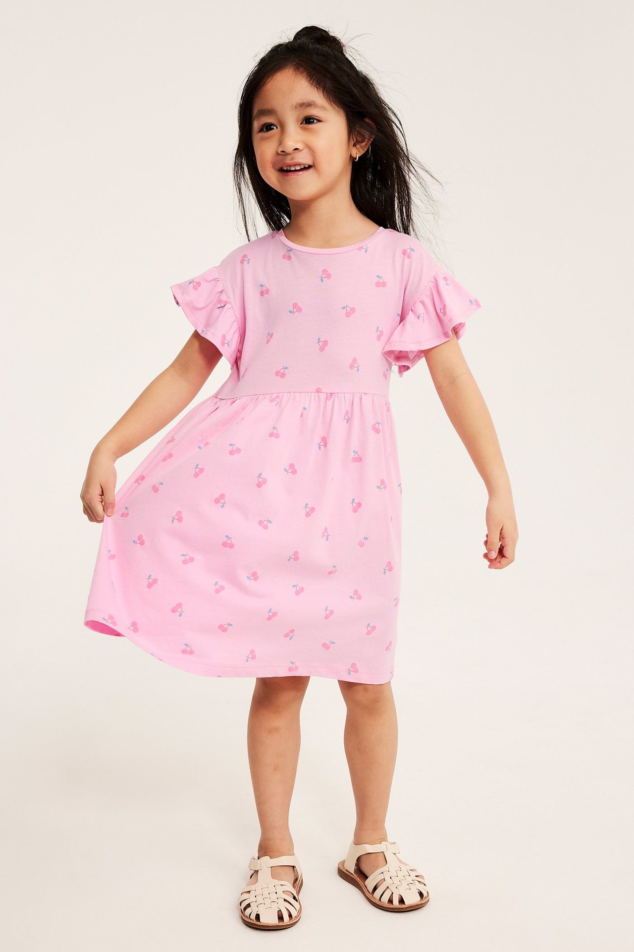 Edestäpäin, lapsi vaaleanpunaisessa puuvillamekossa kirsikkakuviolla, röyhelöhihoilla ja beigenvärisillä sandaaleilla.